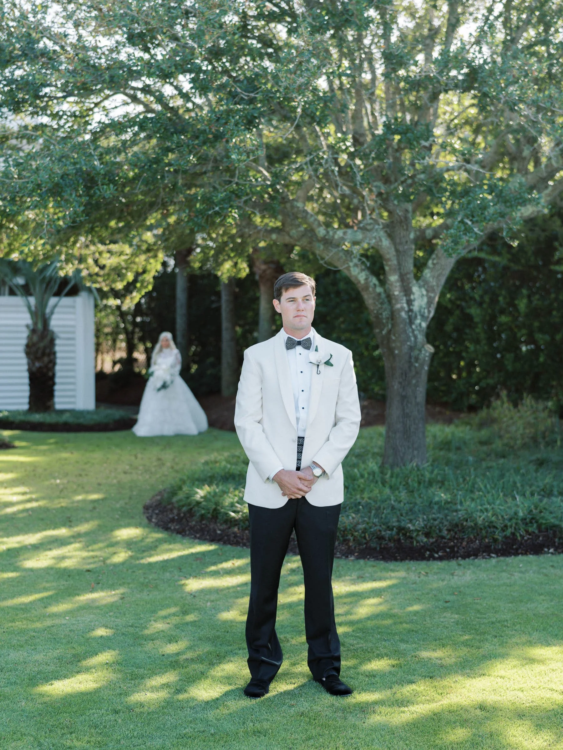 A groom in a white tuxedo jacket and black pants stands with hands clasped, looking serious, outdoors under a large tree. A bride in a white wedding dress with a veil and bouquet stands in the background.