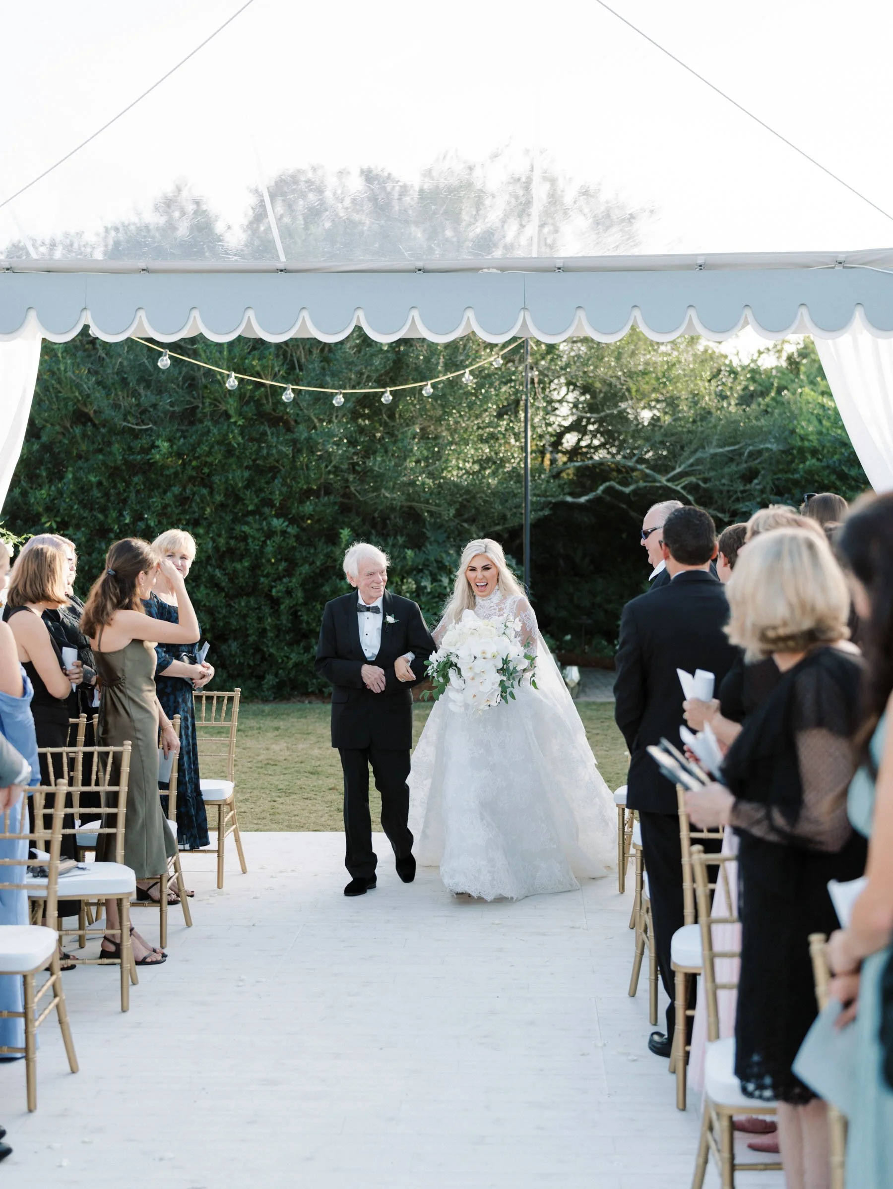 Bride walking down the aisle at an outdoor wedding ceremony, smiling, holding a bouquet, accompanied by her father or officiant, guests seated on both sides, greenery background, under a white tent with string lights.