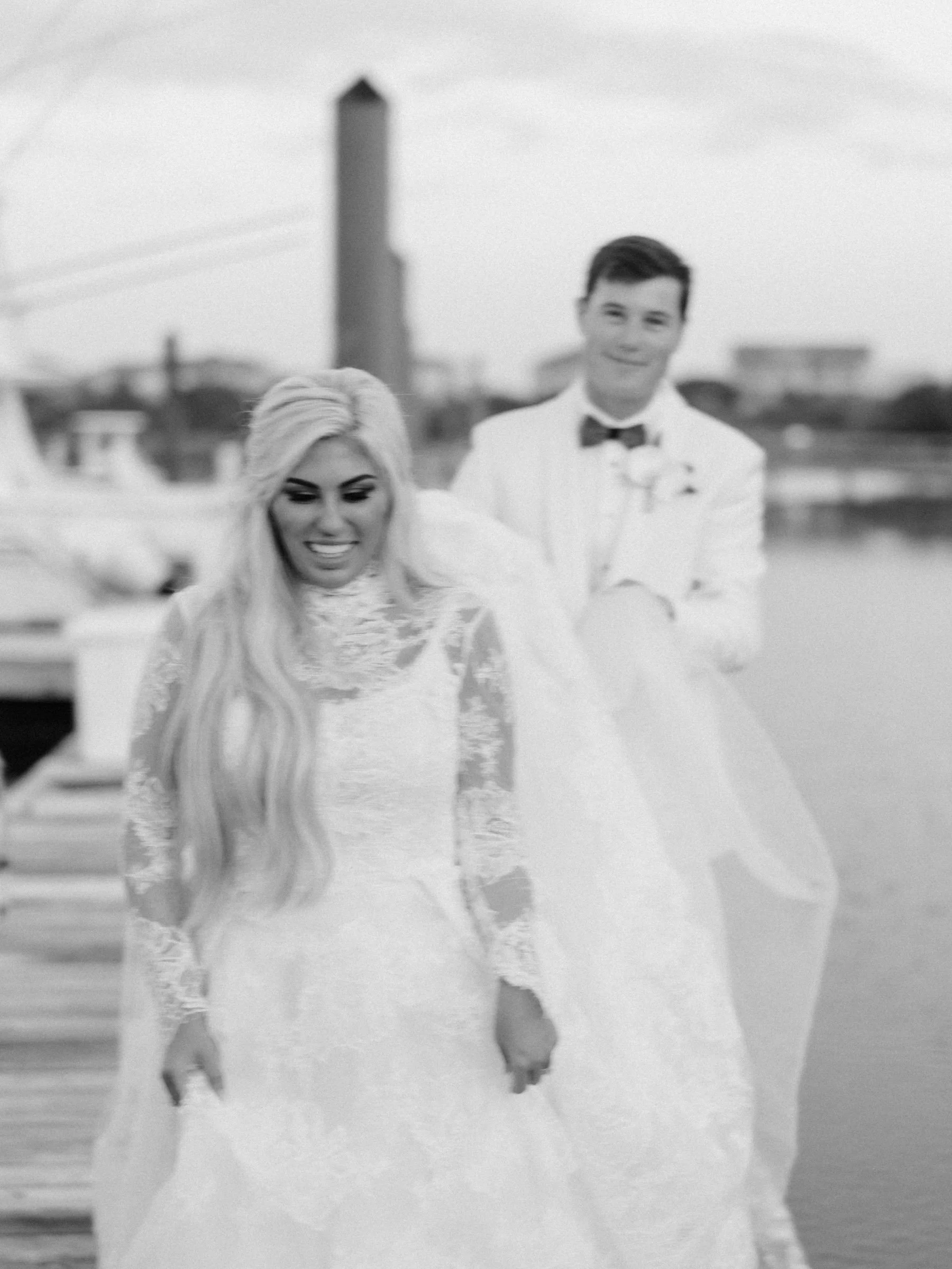 A black and white photo of a bride and groom on a dock near water, with buildings and a bridge in the background. The bride has long, wavy blonde hair and is smiling, wearing a lace wedding dress. The groom, standing slightly behind, has dark hair, a