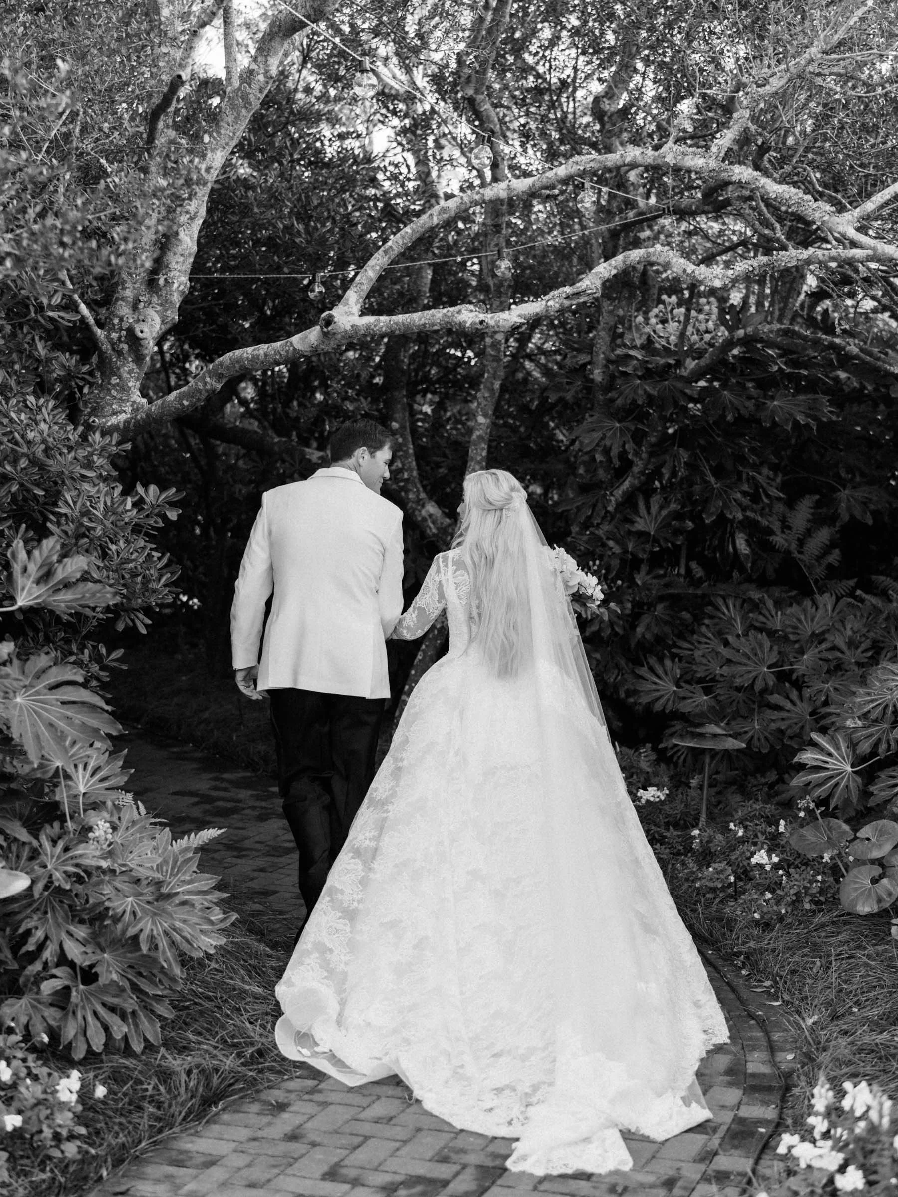 A bride and groom holding hands, walking along a garden path with lush trees and plants, bride in a long lace wedding dress and veil, groom in a light-colored suit jacket.