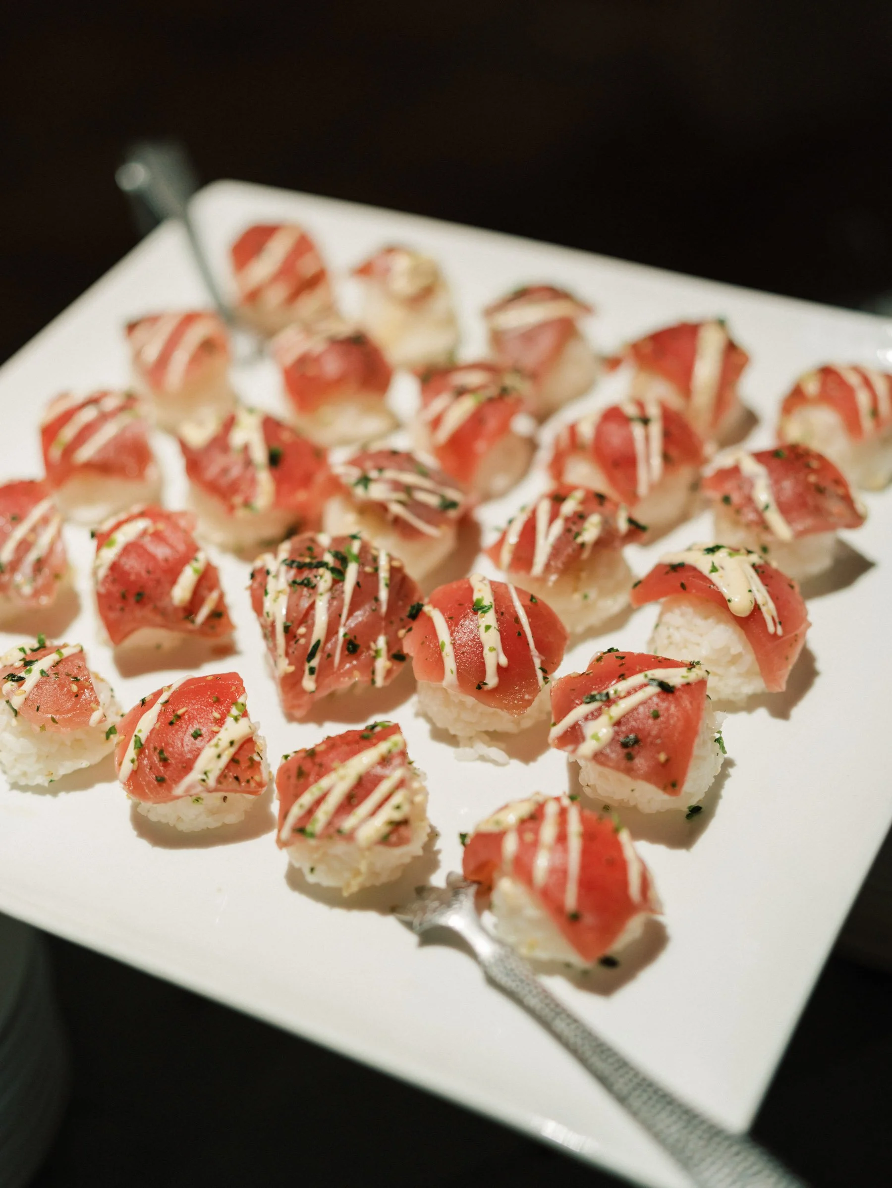 Plate of small sushi bites with slices of raw fish drizzled with sauce and garnished with herbs, on a white square platter.