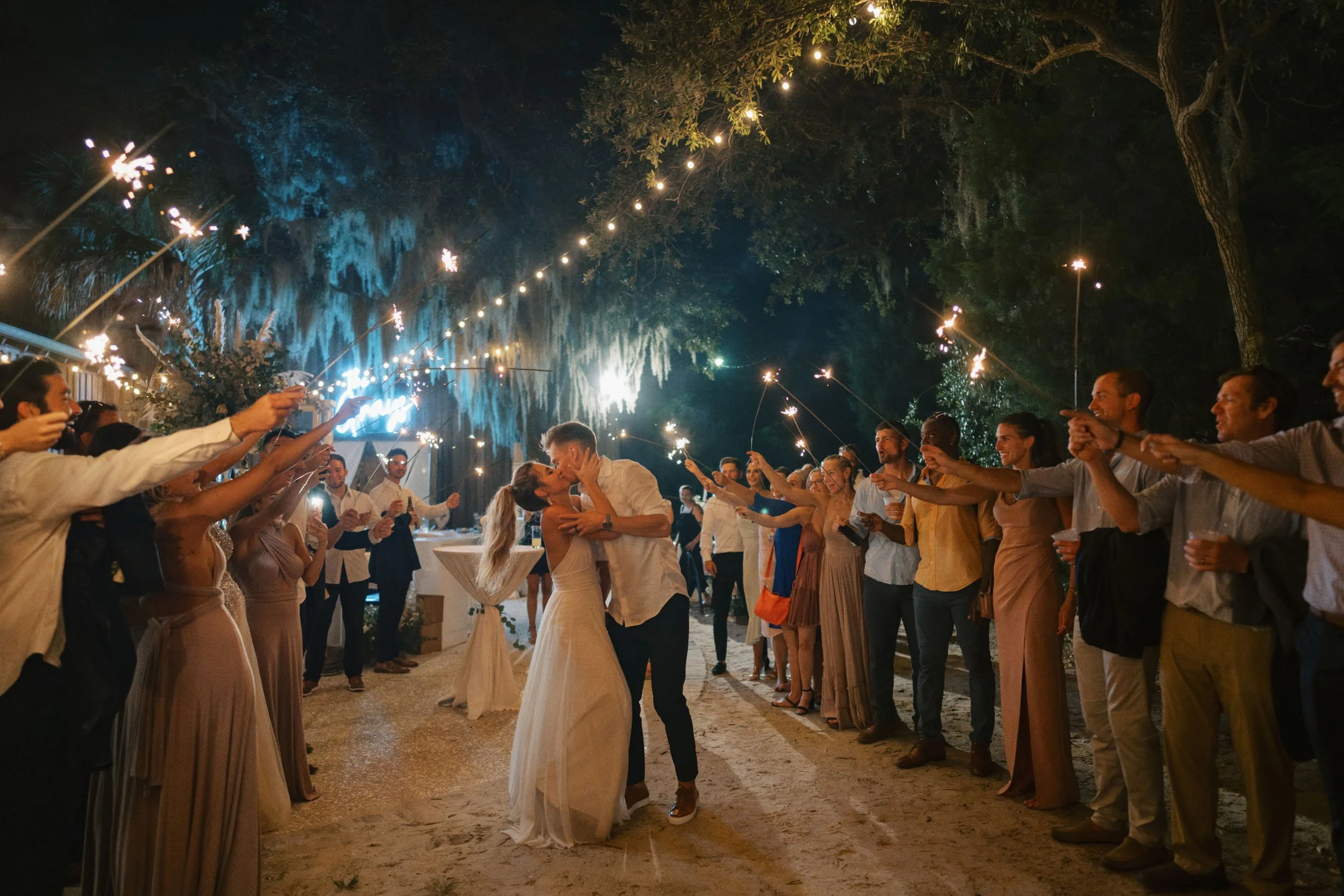 A bride and groom kiss surrounded by guests holding sparklers during an outdoor evening celebration at a wedding reception.