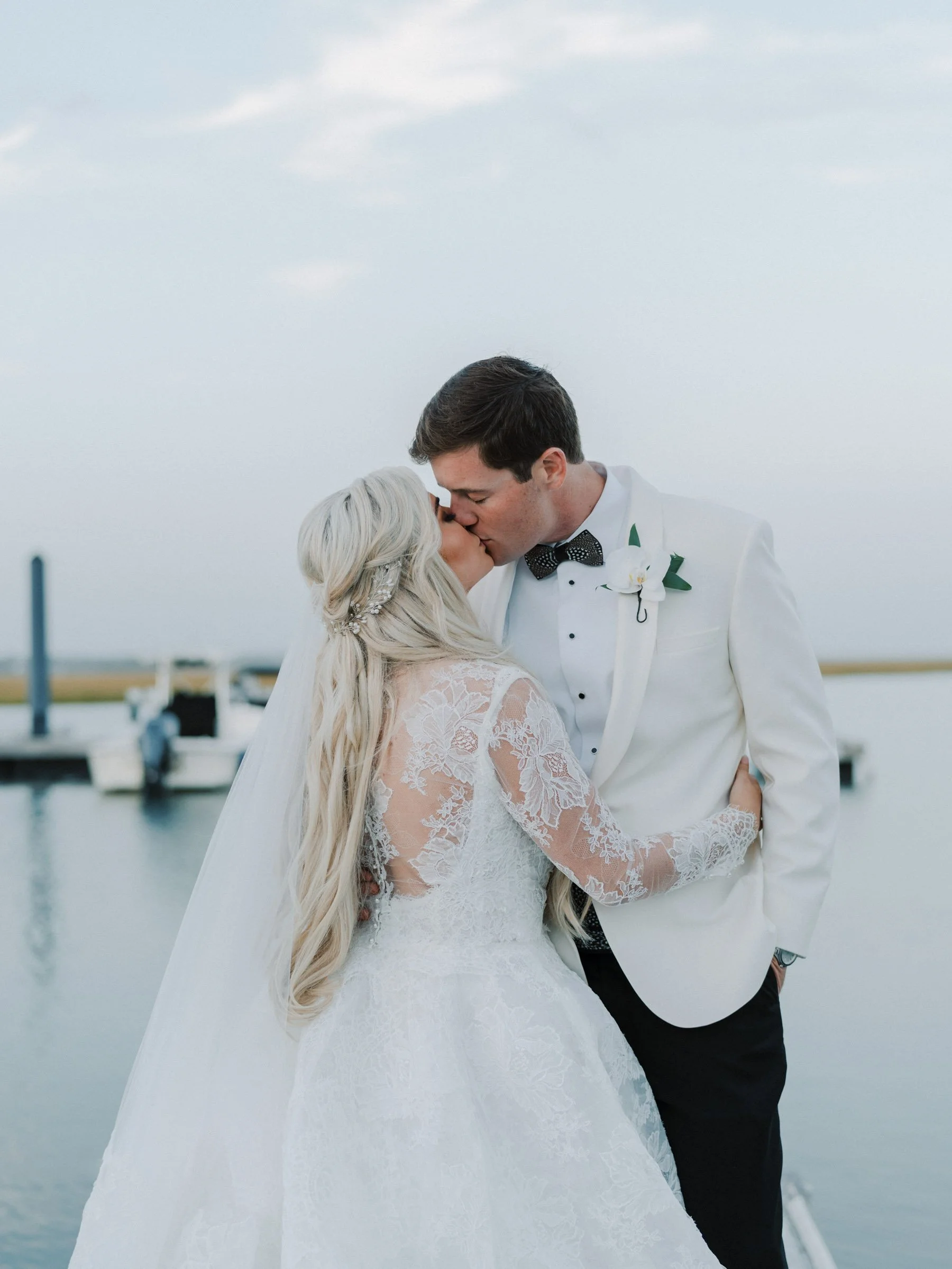A couple in wedding attire sharing a kiss by the water at sunset, with docks and boats in the background.