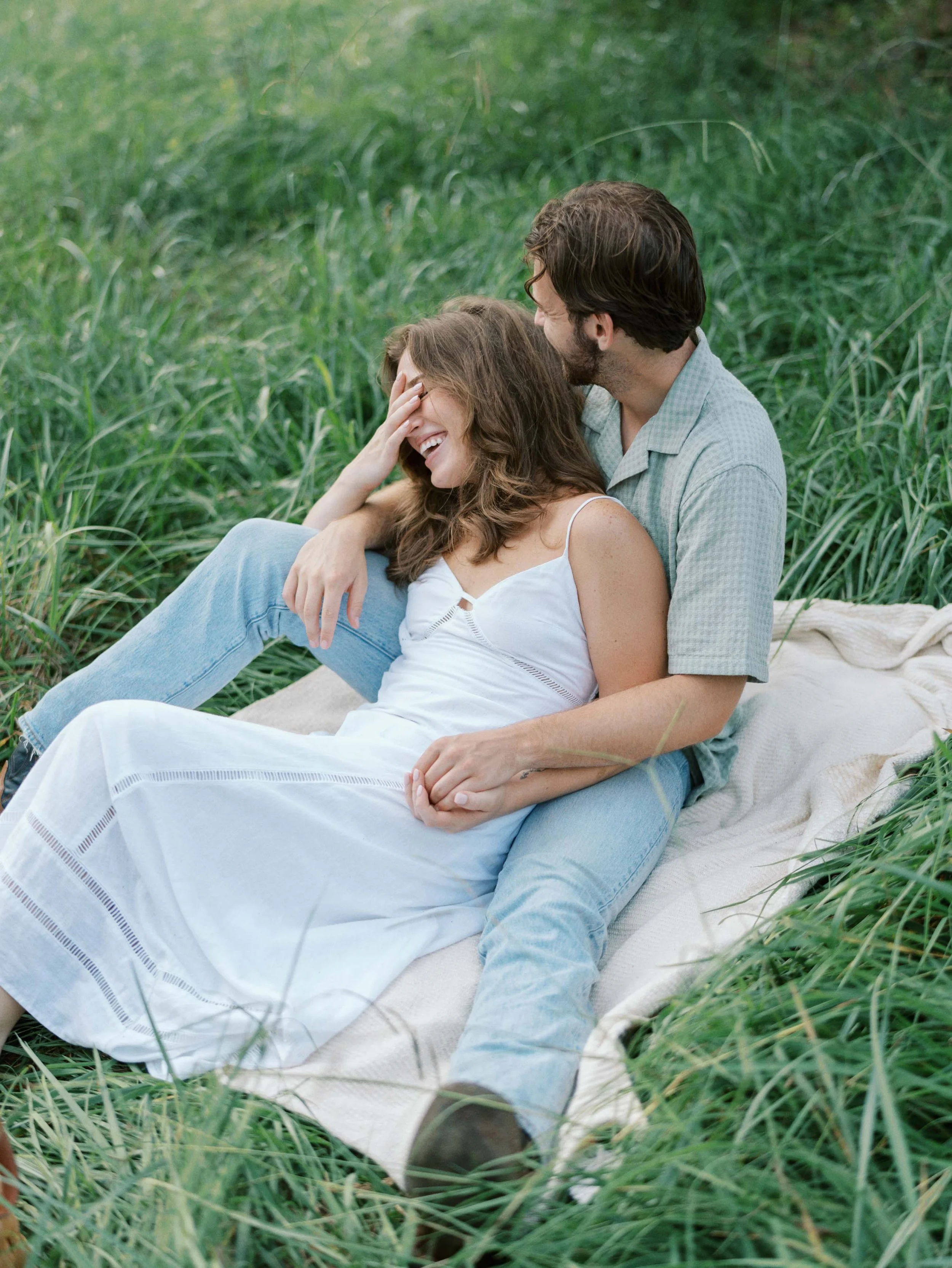 A couple sitting on a blanket in a grassy field, laughing together with one person touching their face.