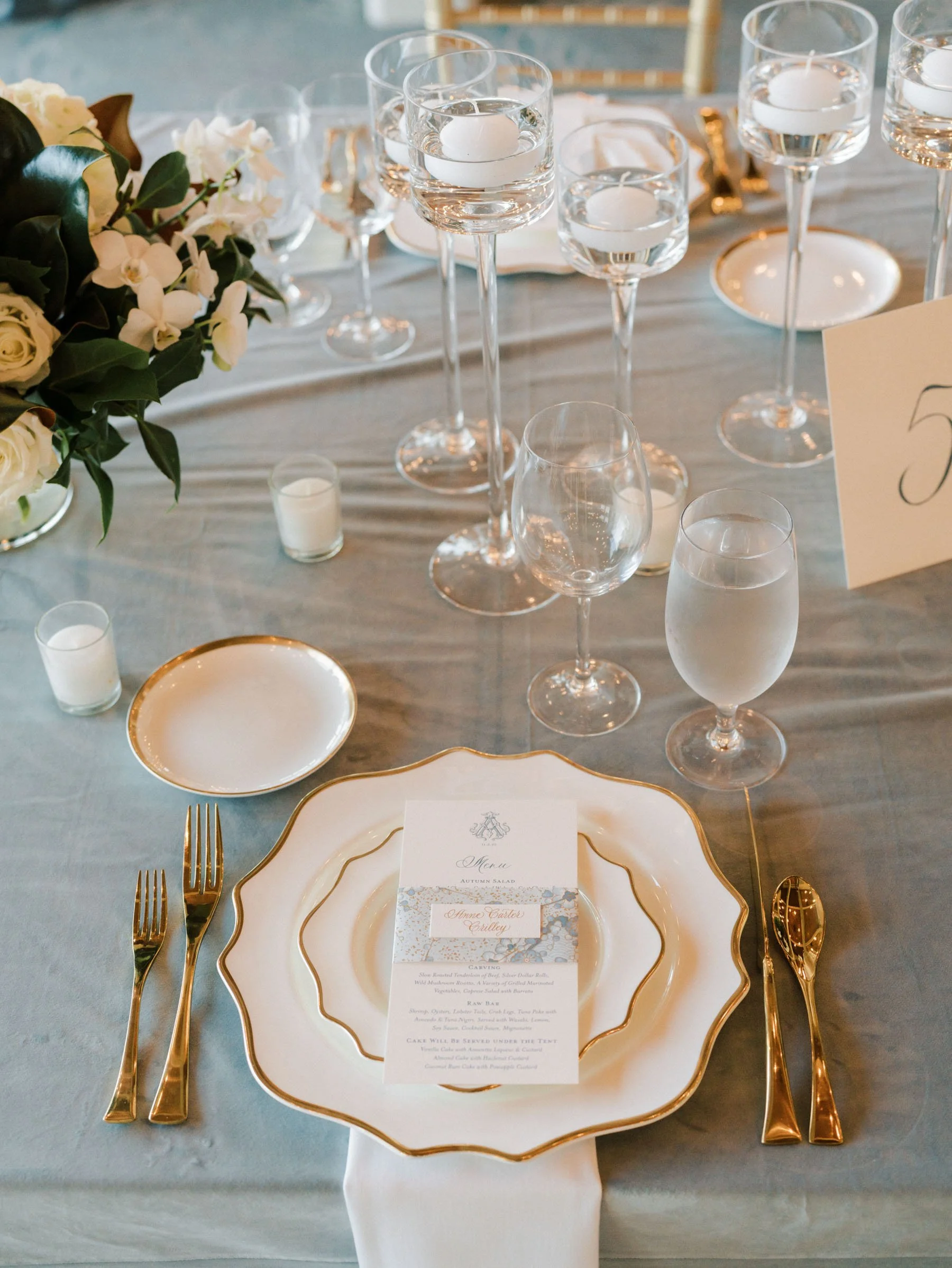 Elegant table setting with white and gold-rimmed plates, gold flatware, water glasses, and floating candles. A floral centerpiece with white and green flowers. A menu card on the plate.