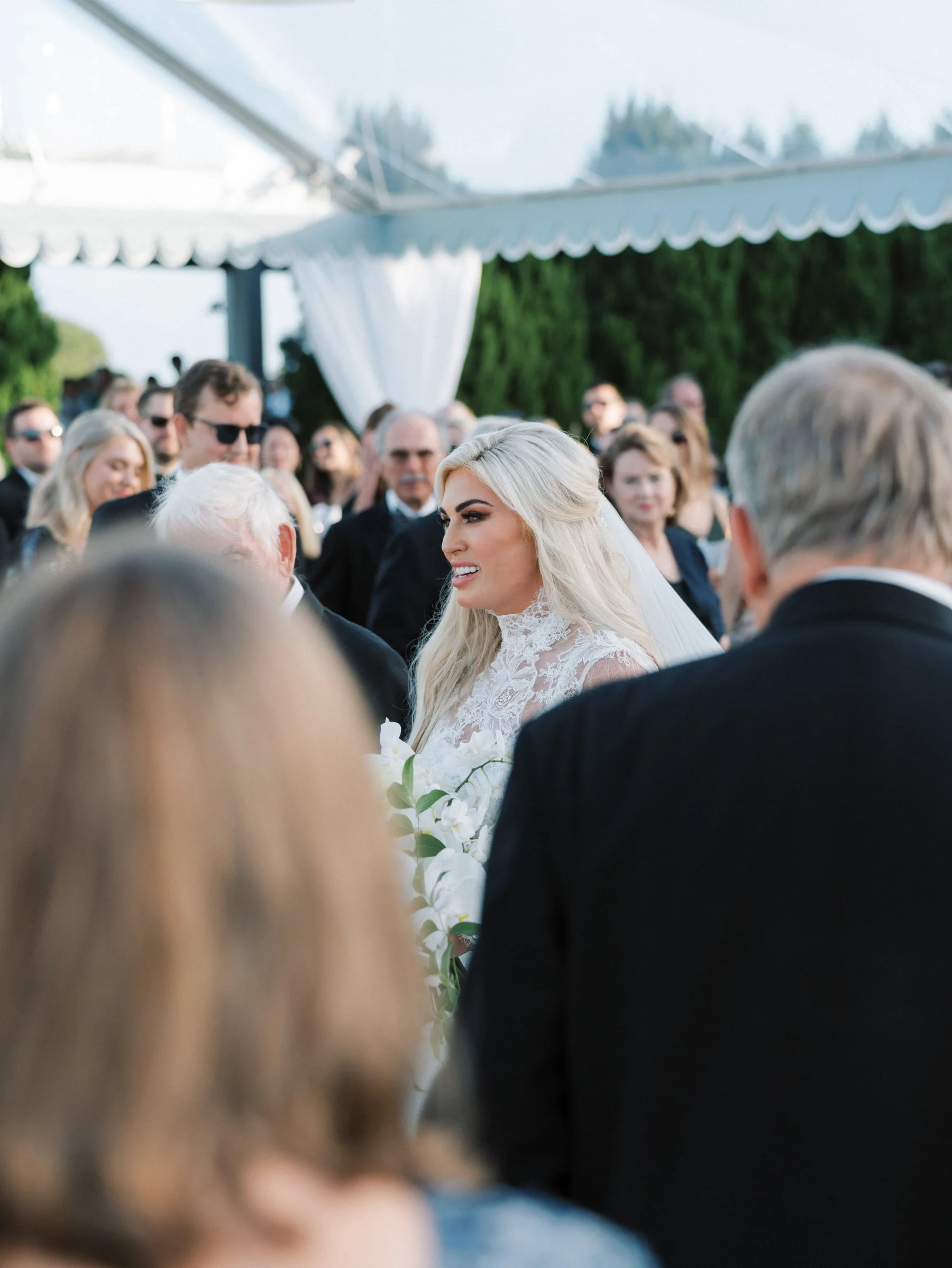 Bride with long blonde hair and lace wedding dress smiling during outdoor wedding ceremony surrounded by guests under a canopy.