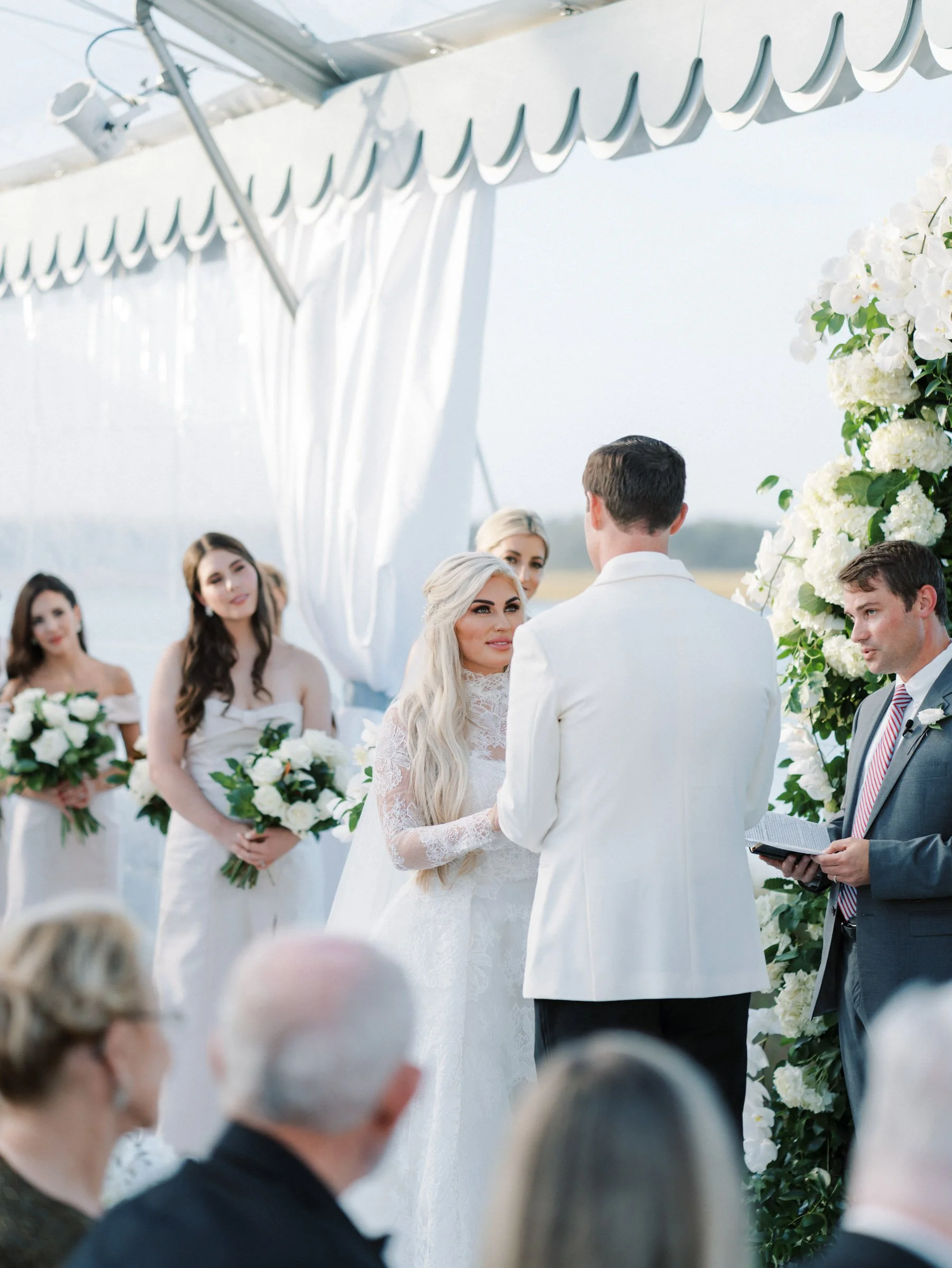 Bride and groom exchanging wedding vows under an outdoor white canopy decorated with flowers, with bridesmaids holding bouquets in the background.