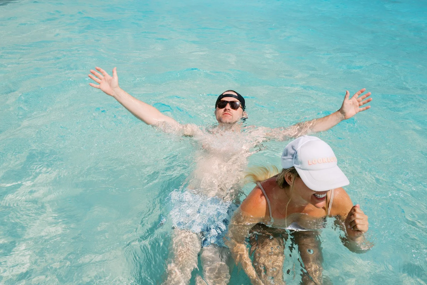 Two people in a swimming pool enjoying the water, with one person swimming and the other woman smiling under a white cap.
