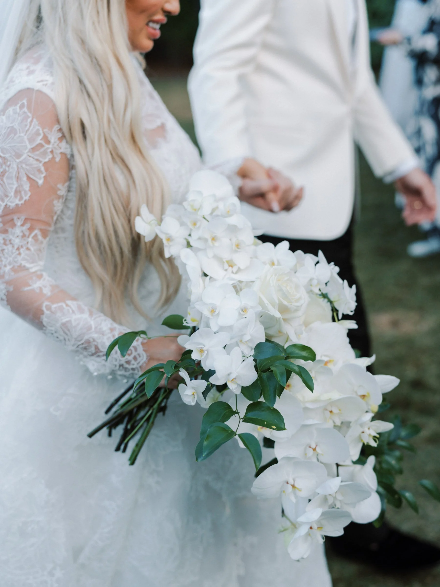 A bride holding a cascading bouquet of white flowers, including orchids and roses, during a wedding ceremony, with her groom holding her hand.