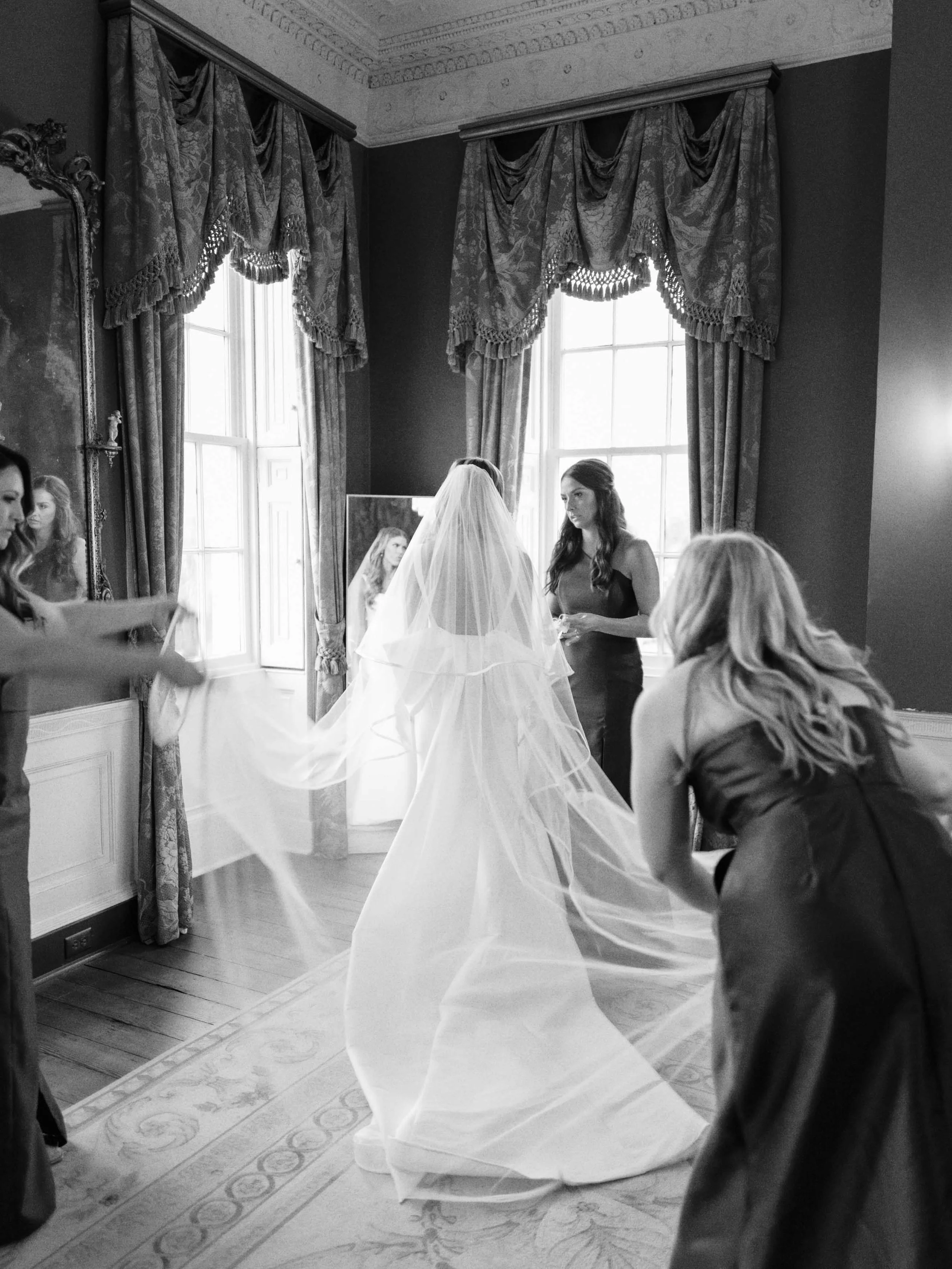A bride walking in a wedding dress with veil as her bridesmaids help her prepare in a decorated room with large windows, ornate curtains, and a mirror.