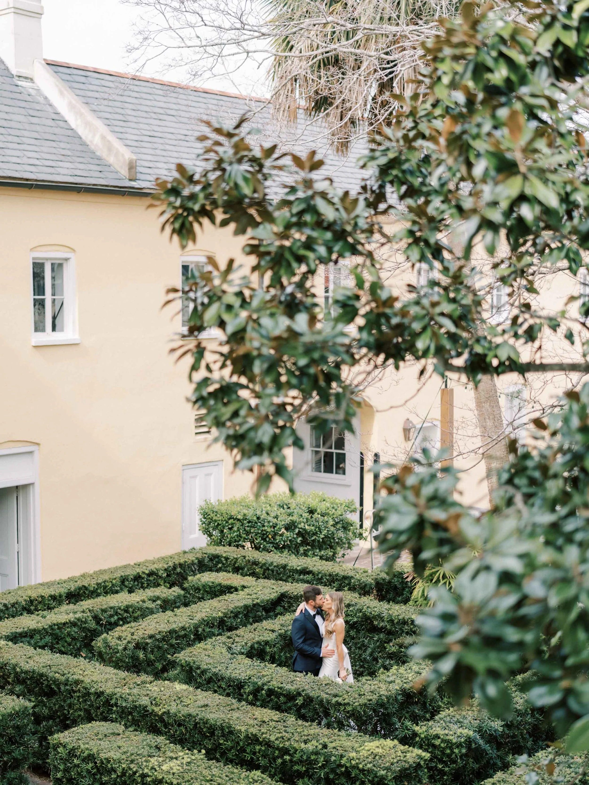 A couple in wedding attire kissing in a hedge maze garden outside a yellow house.
