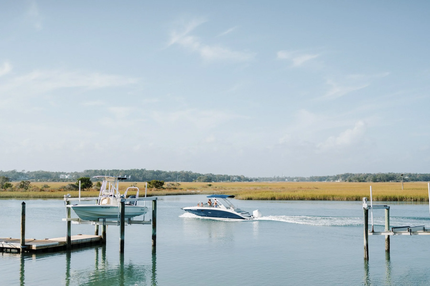A boat dock on a body of water with a boat moving across, leaving a wake behind it. In the background, there is marshland and trees under a partly cloudy sky.
