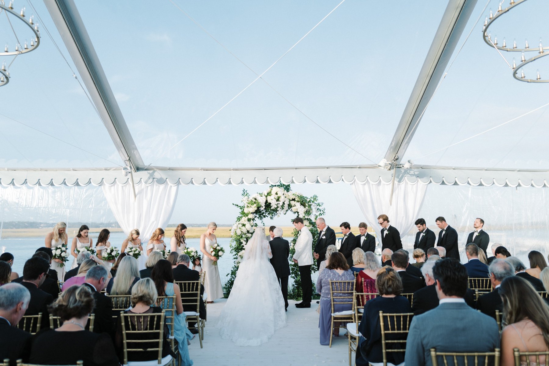 A wedding ceremony taking place under a large white tent with water and open landscape in the background. The bride and groom stand beneath an arch decorated with white flowers and greenery. Bridesmaids and groomsmen stand beside them, with guests se