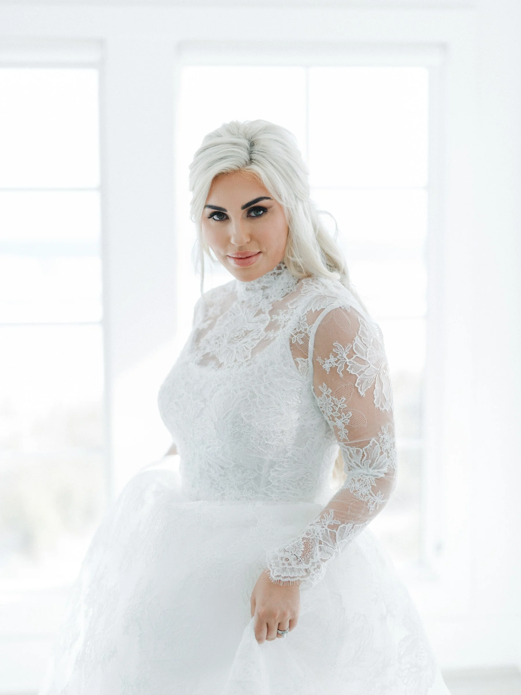A woman with platinum blonde hair in a wedding dress standing in front of large windows.