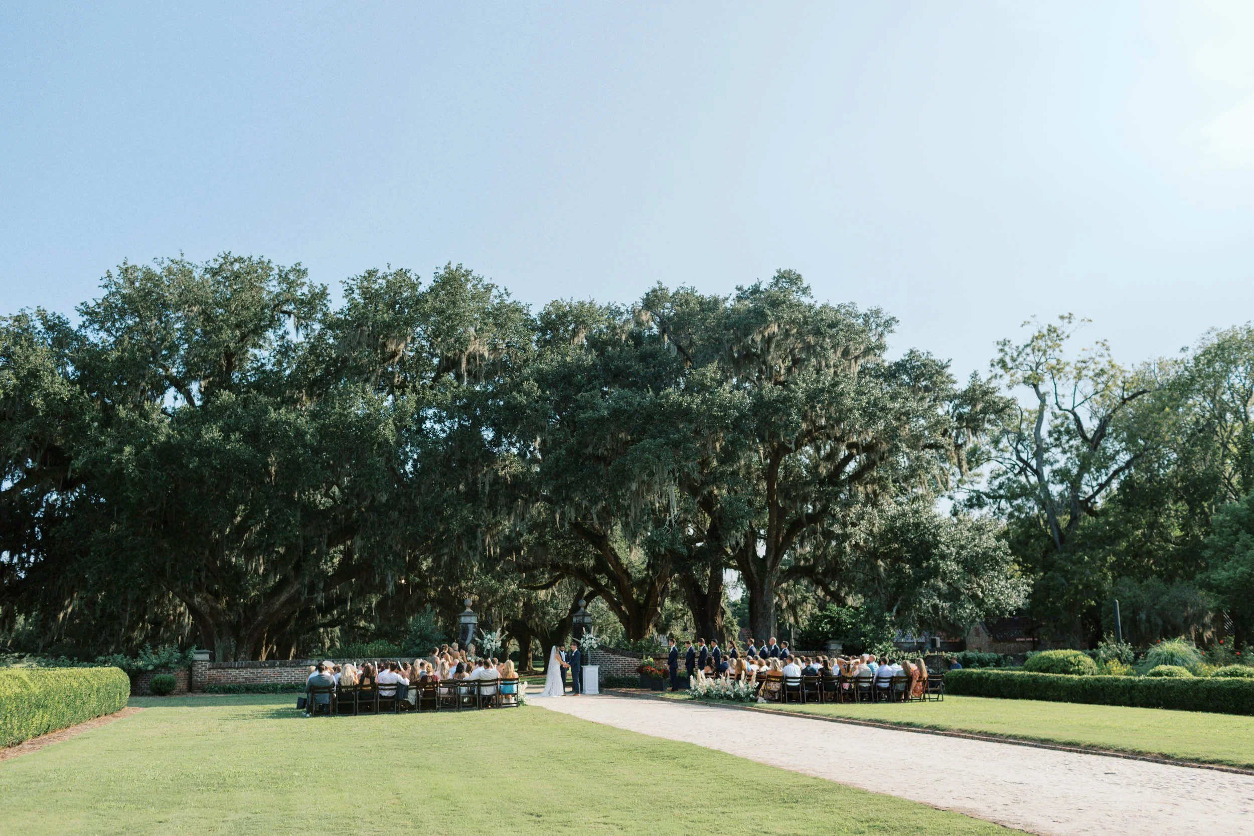 Wedding ceremony outdoors under large oak trees on a sunny day with guests seated on either side of a white aisle at Boone Hall.