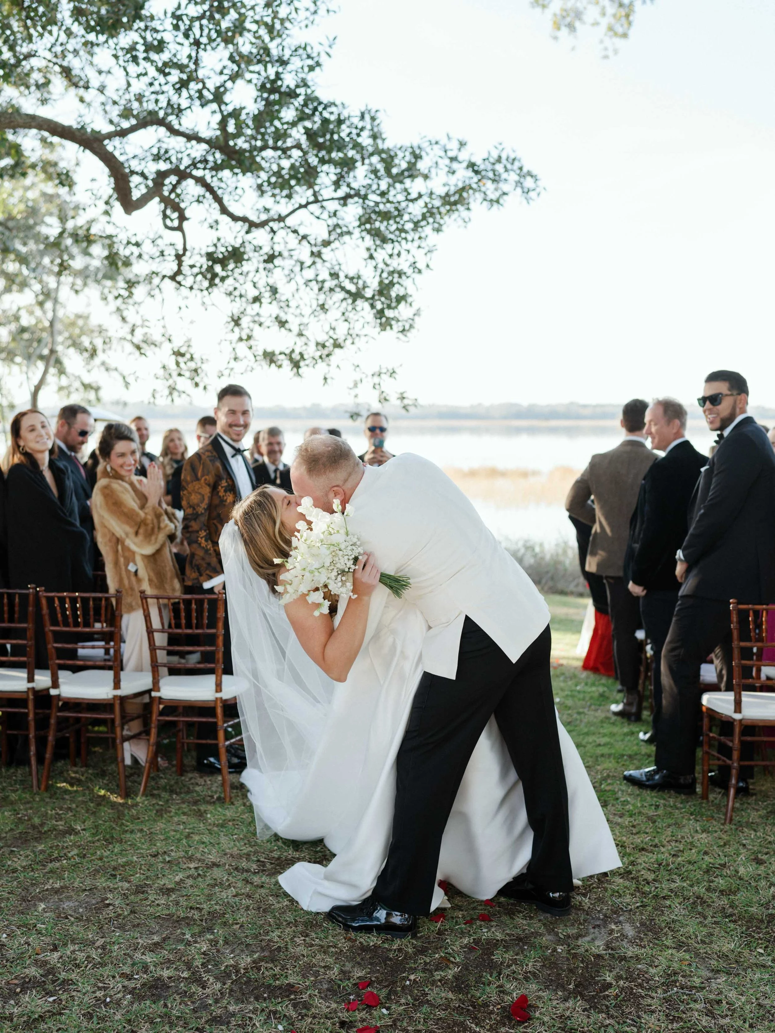 A newlywed couple sharing a kiss during their outdoor wedding ceremony by a lake, surrounded by friends and family, with trees and a lake in the background.