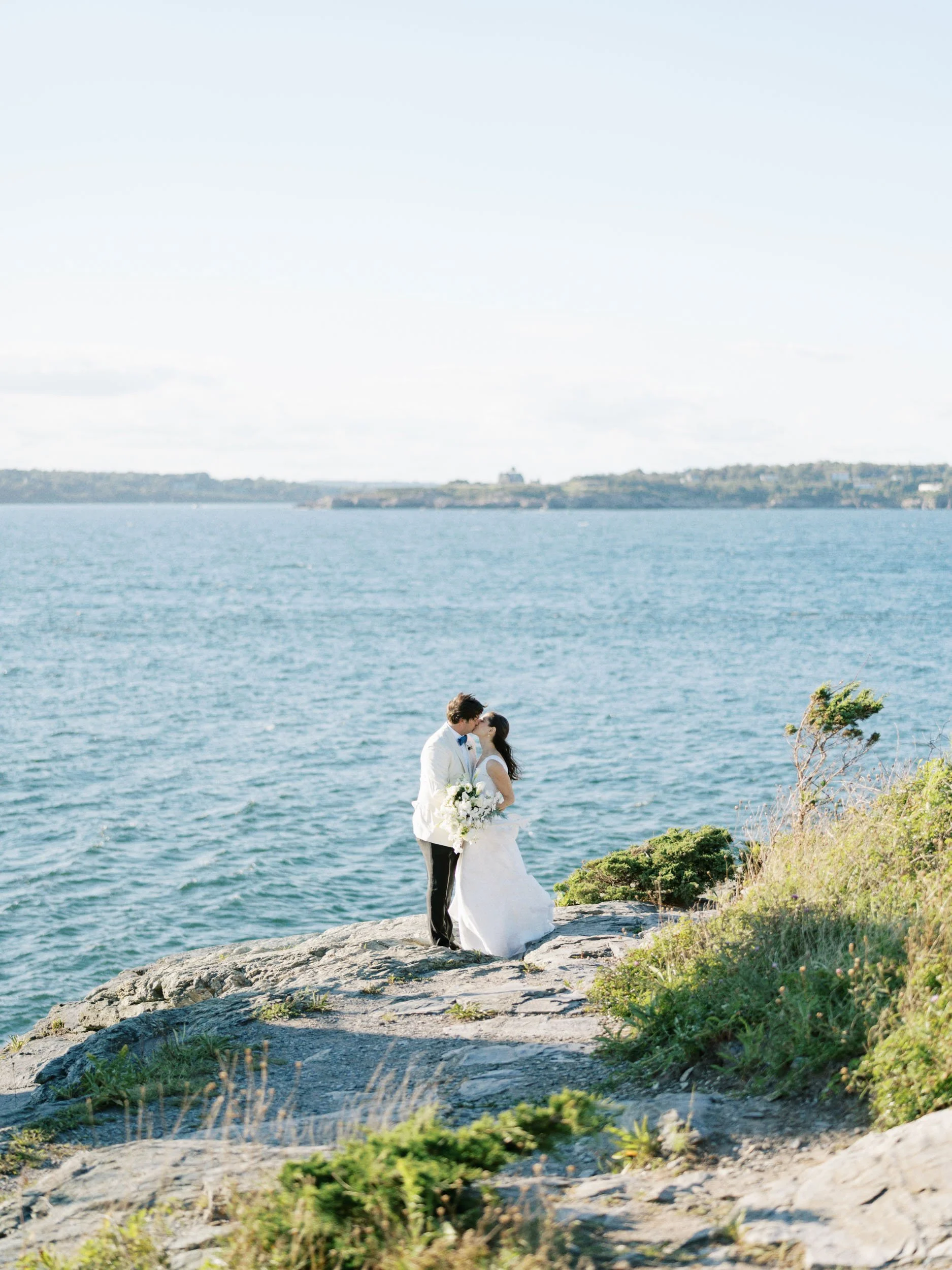 A couple in wedding attire sharing a kiss on a rocky cliff by the water at Castle Hill, with greenery and the ocean in the background.