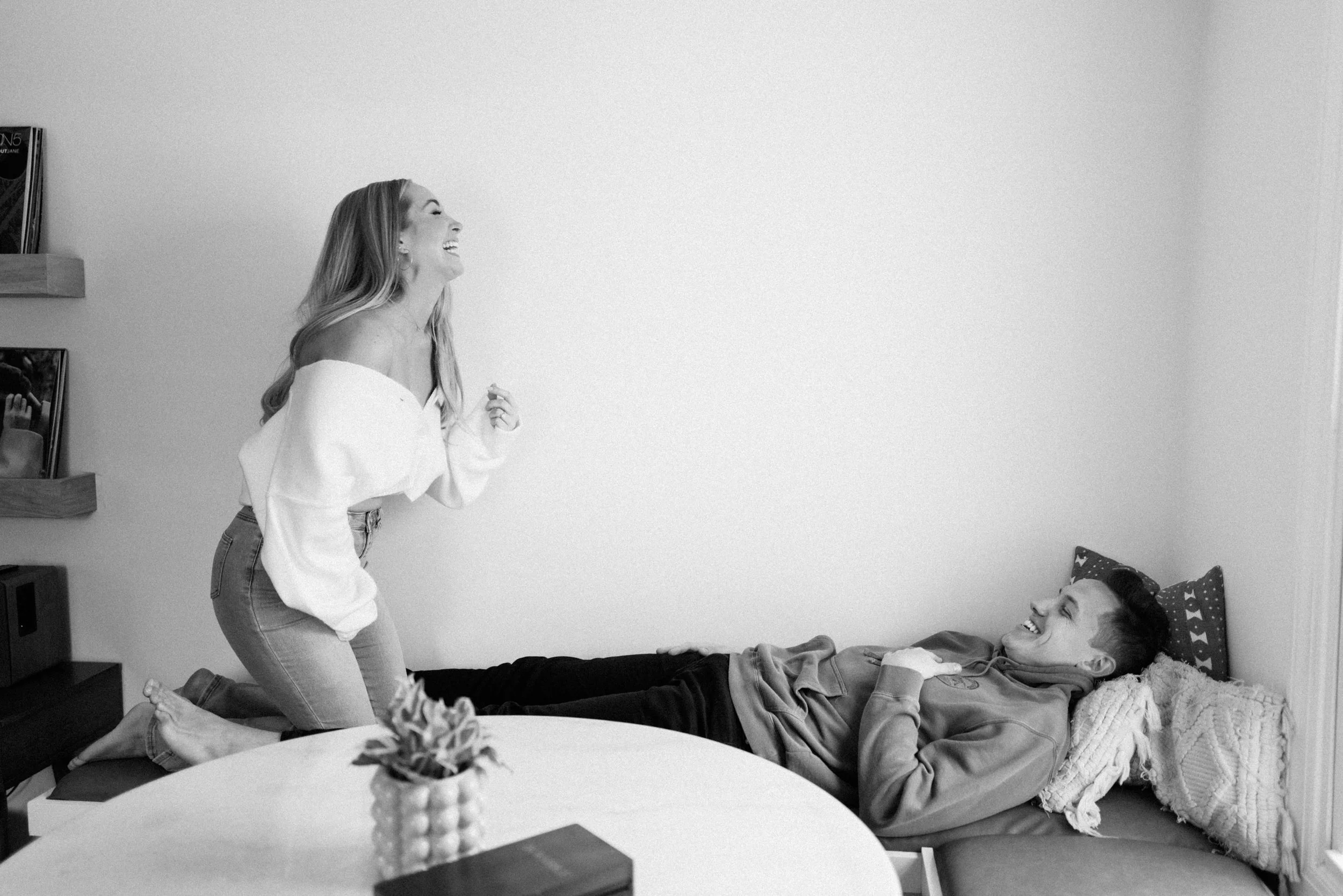 A woman laughing and leaning forward towards a man lying on a couch, also laughing. The woman is standing on the couch with her knees bent, wearing an off-the-shoulder top and jeans. The man is relaxed with his head on pillows, wearing a jacket. A ta