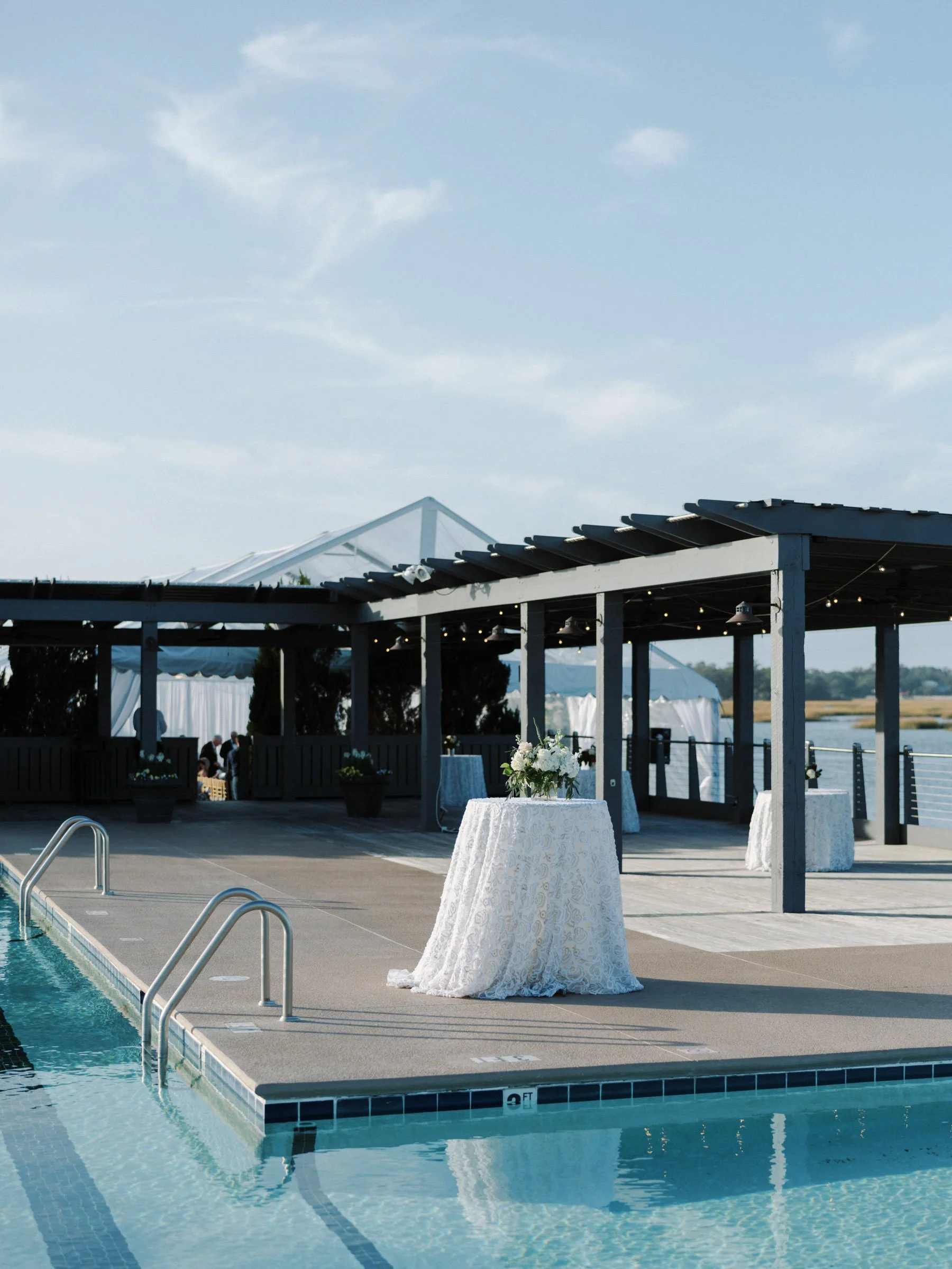 A decorated outdoor area near a swimming pool with tables draped in white tablecloths and floral centerpieces, under a wooden pergola with string lights, set up for a special event.