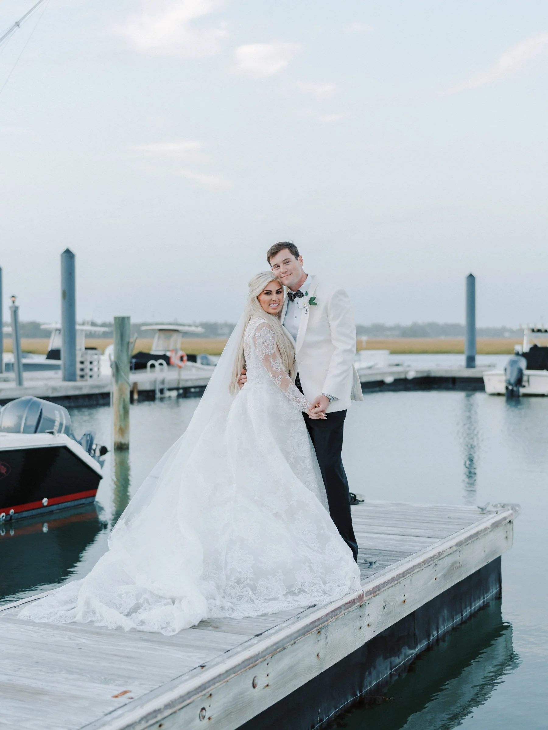 A newlywed couple in wedding attire standing on a dock by the water, smiling at the camera, with boats in the background.