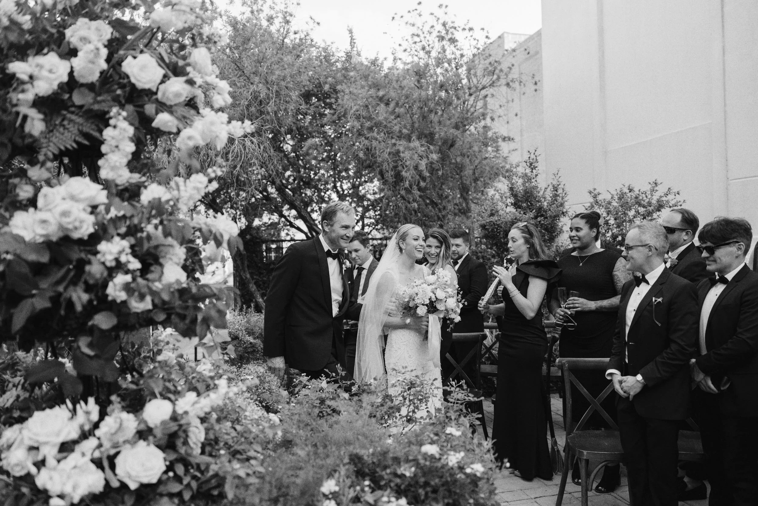 A black and white photo of a wedding ceremony outdoors with the bride and groom smiling, surrounded by friends and family, with floral decorations and trees in the background.