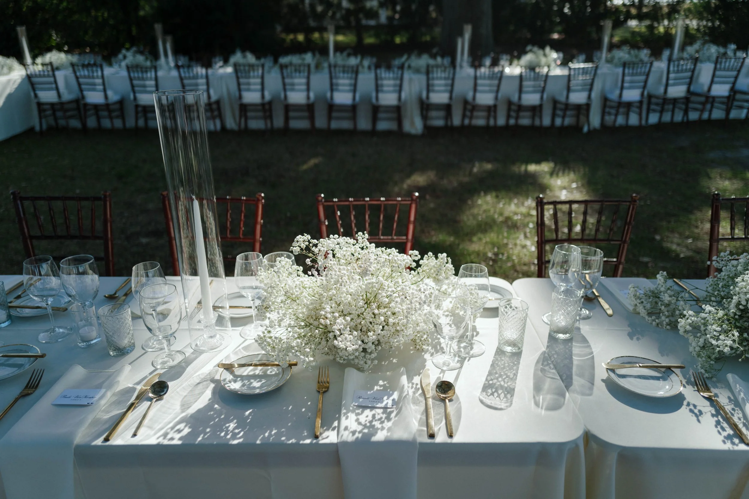 Elegant outdoor dinner setup with white tablecloth, floral arrangement, glassware, and gold utensils, surrounded by wooden chairs, on a grassy area at The Riverhouse at Lowndes Grove in Charleston, SC. 