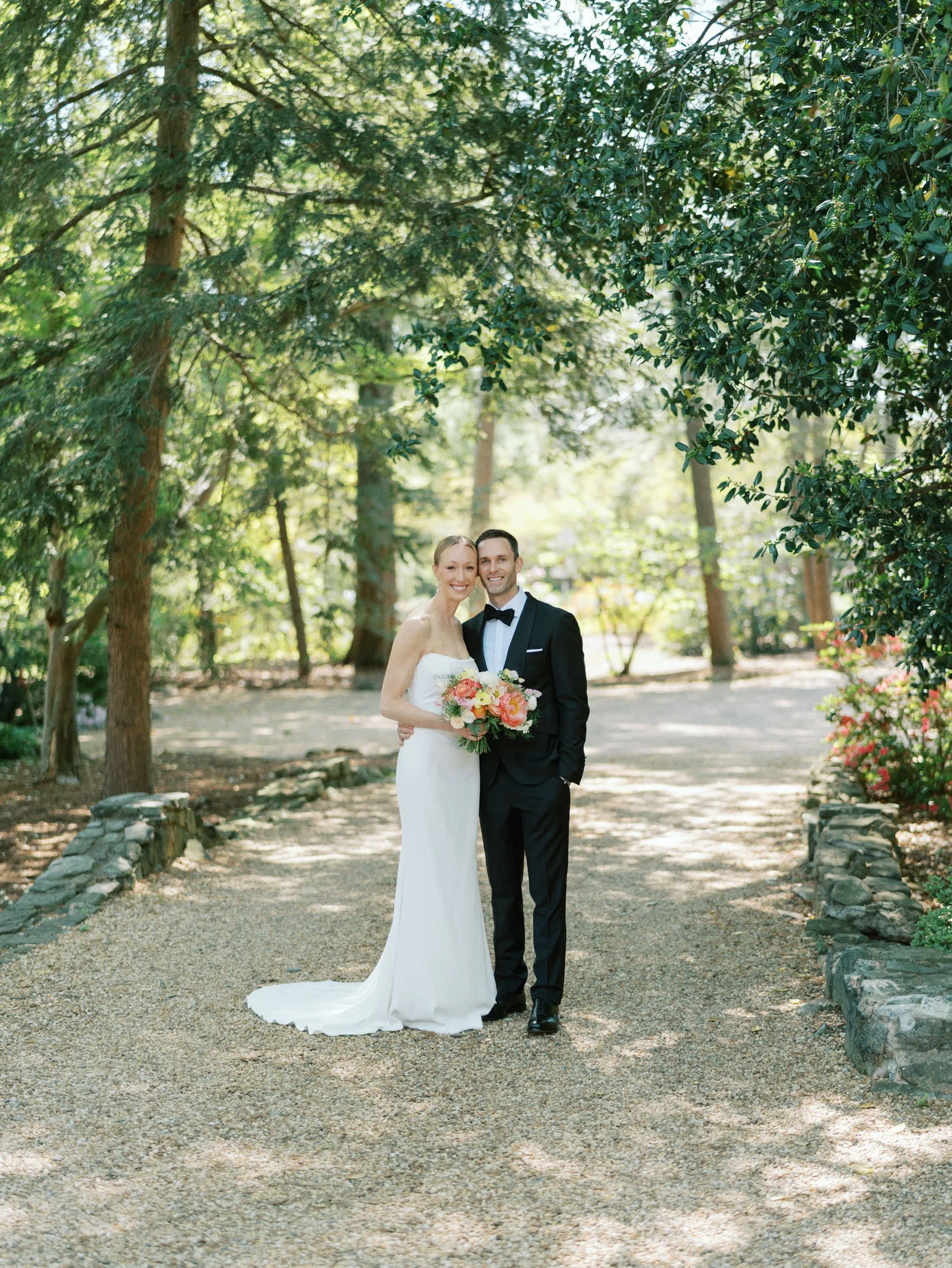 A bride and groom standing together in a forested area at Vanlandingham Estate in Charlotte, dressed in wedding attire. The bride is in a white gown holding a bouquet of flowers, and the groom is in a black tuxedo.