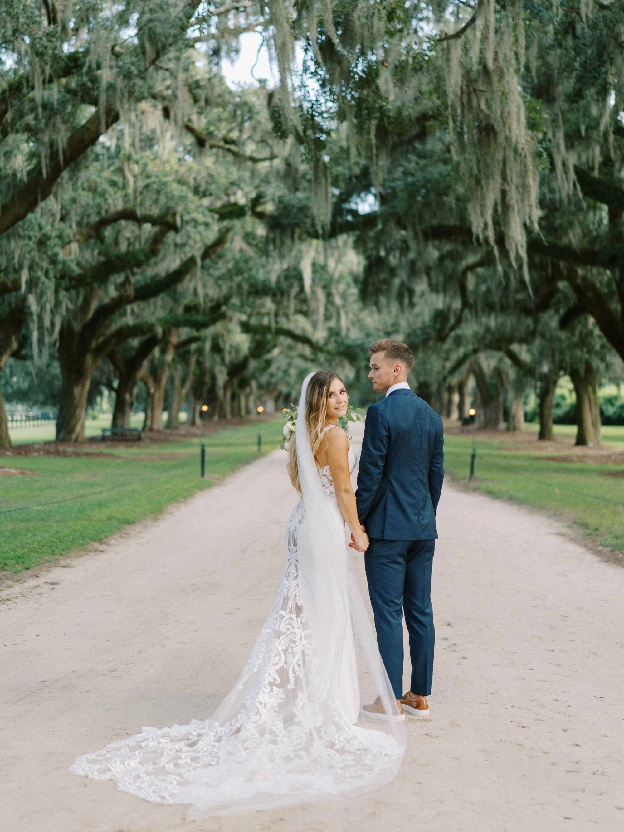 A bride and groom holding hands and looking at each other in a tree-lined park.