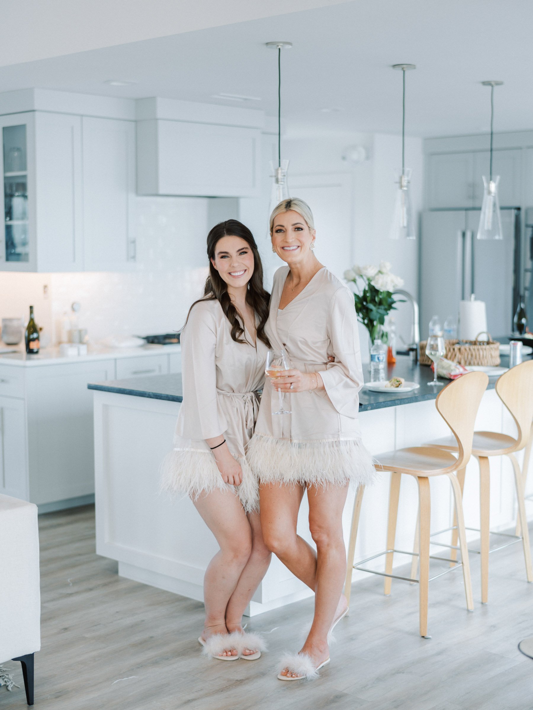 Two women in beige pajamas with feather trim standing in a modern, white kitchen, smiling, holding wine glasses, with bar stools and kitchen appliances in the background.