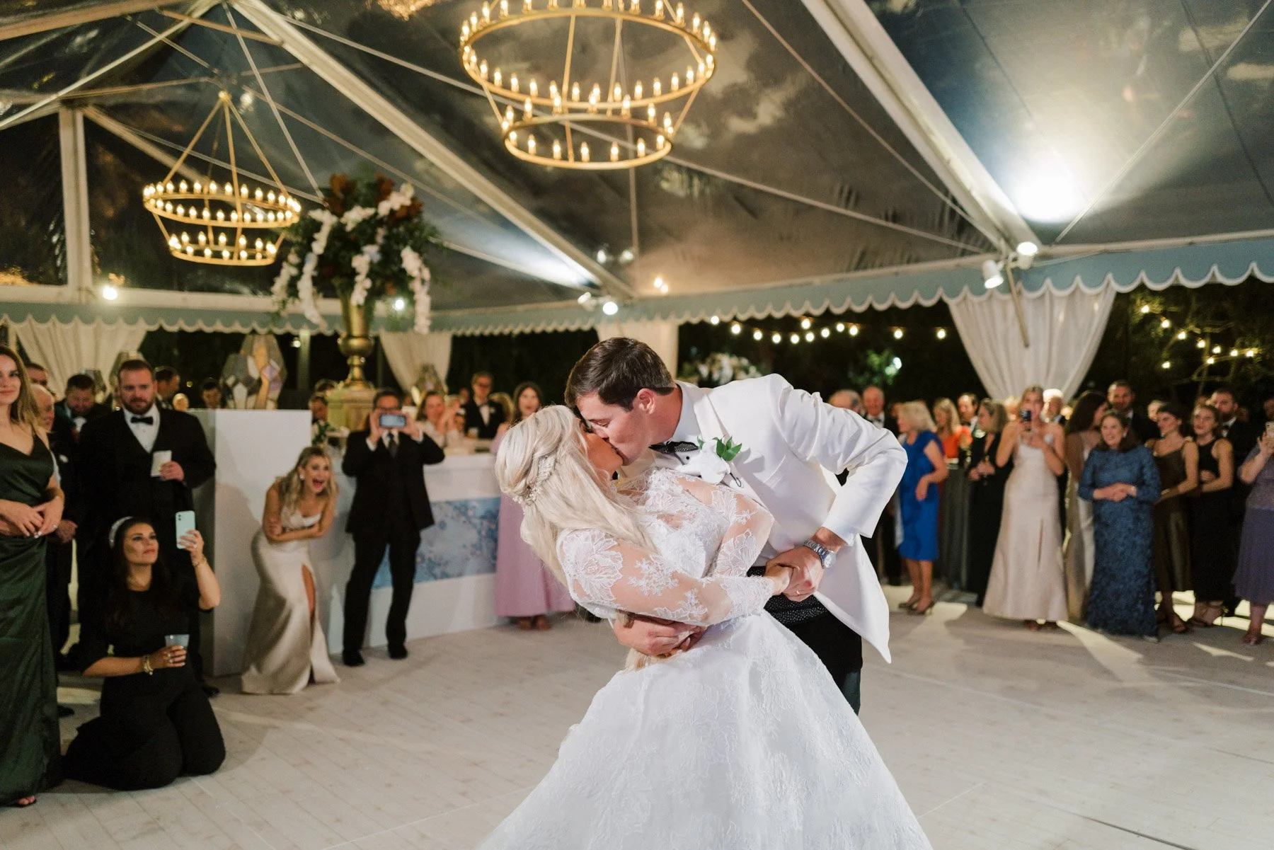 A bride and groom sharing a first dance at their wedding reception, surrounded by guests under a large decorated tent with hanging chandeliers.