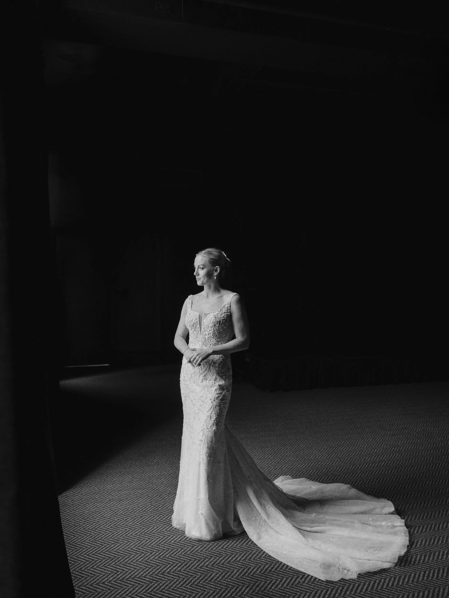 A woman in a wedding dress standing alone in a dark room with spotlight focusing on her.