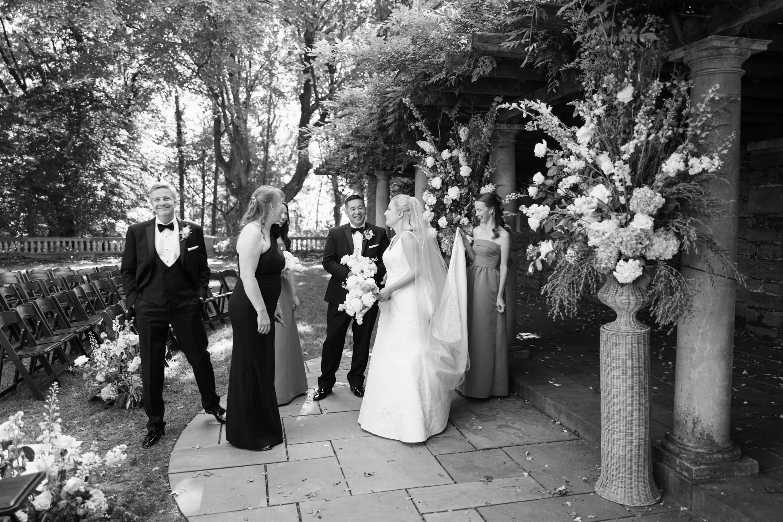 A black-and-white photograph of a wedding ceremony outdoors featuring the bride in a white gown and veil, holding a bouquet, standing with the groom in a tuxedo. The officiant and three women in dresses are also present, with chairs and floral arrangements in the background surrounded by trees.