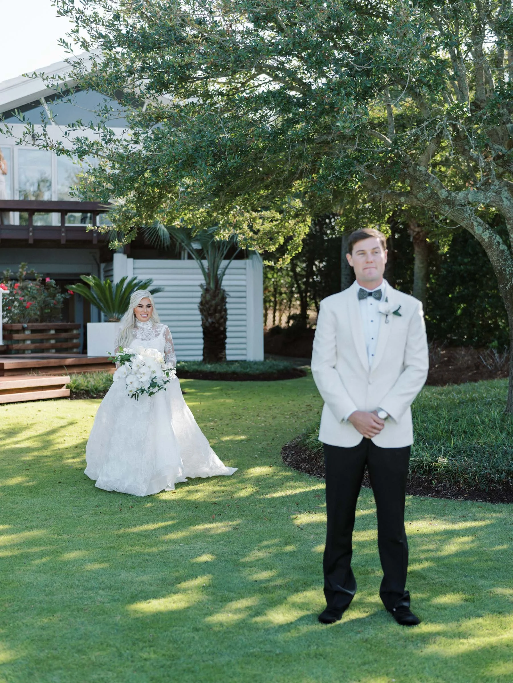 A bride in a white lace wedding dress holding a bouquet of white flowers stands in the background, smiling. A groom in a white tuxedo jacket, black pants, and bow tie stands in the foreground with his hands clasped, outdoors in a garden with trees an