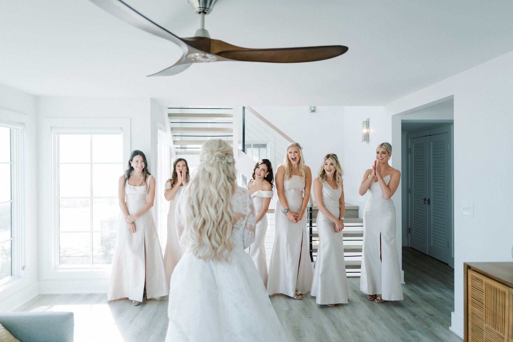 A bride with long, curly blonde hair facing six bridesmaids, all in white dresses, inside a bright, modern room with large windows and wooden flooring.