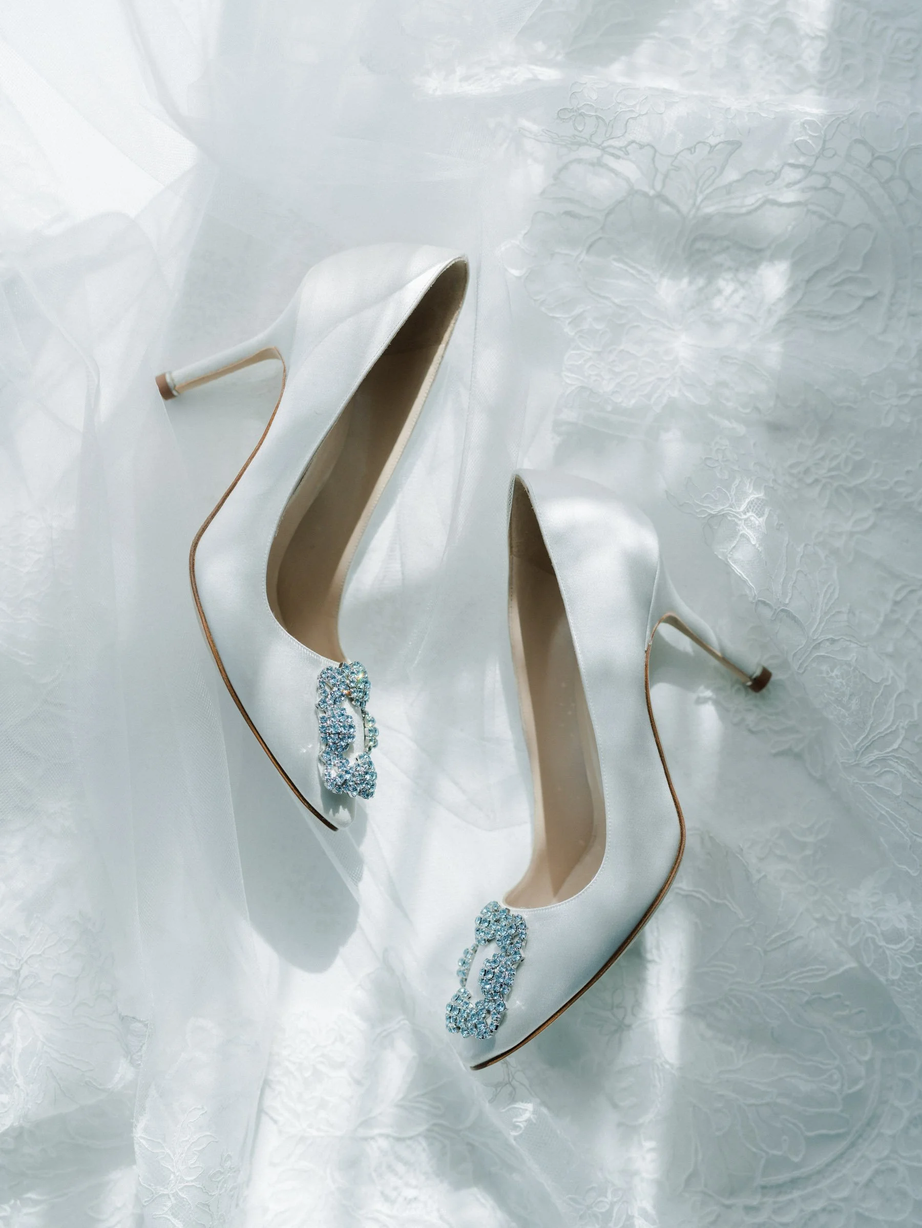 A pair of elegant white satin high-heeled shoes with jeweled embellishments on the toes, placed on a white textured fabric background.