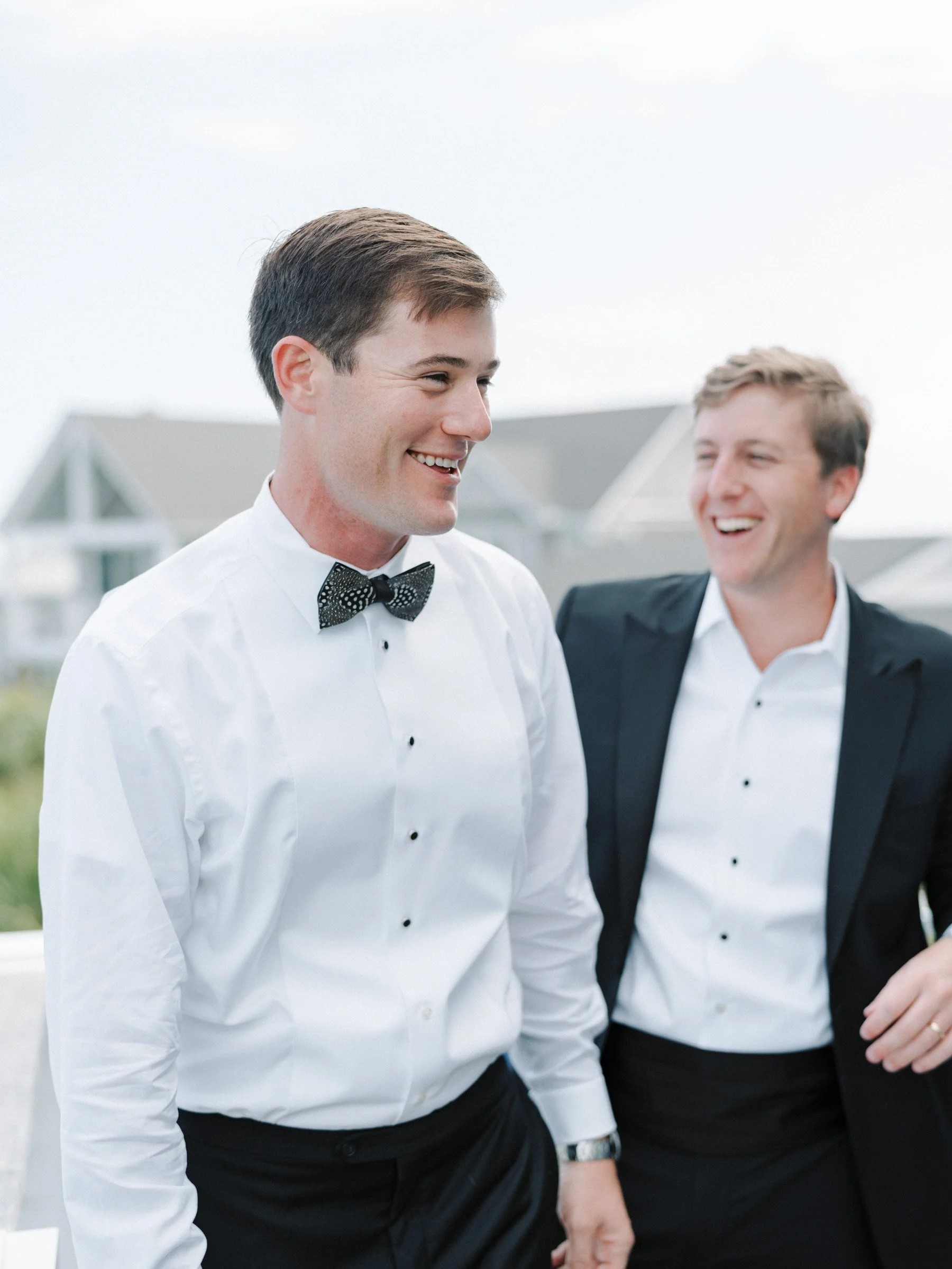 Two men in tuxedos laughing and smiling at each other outdoors with houses in the background