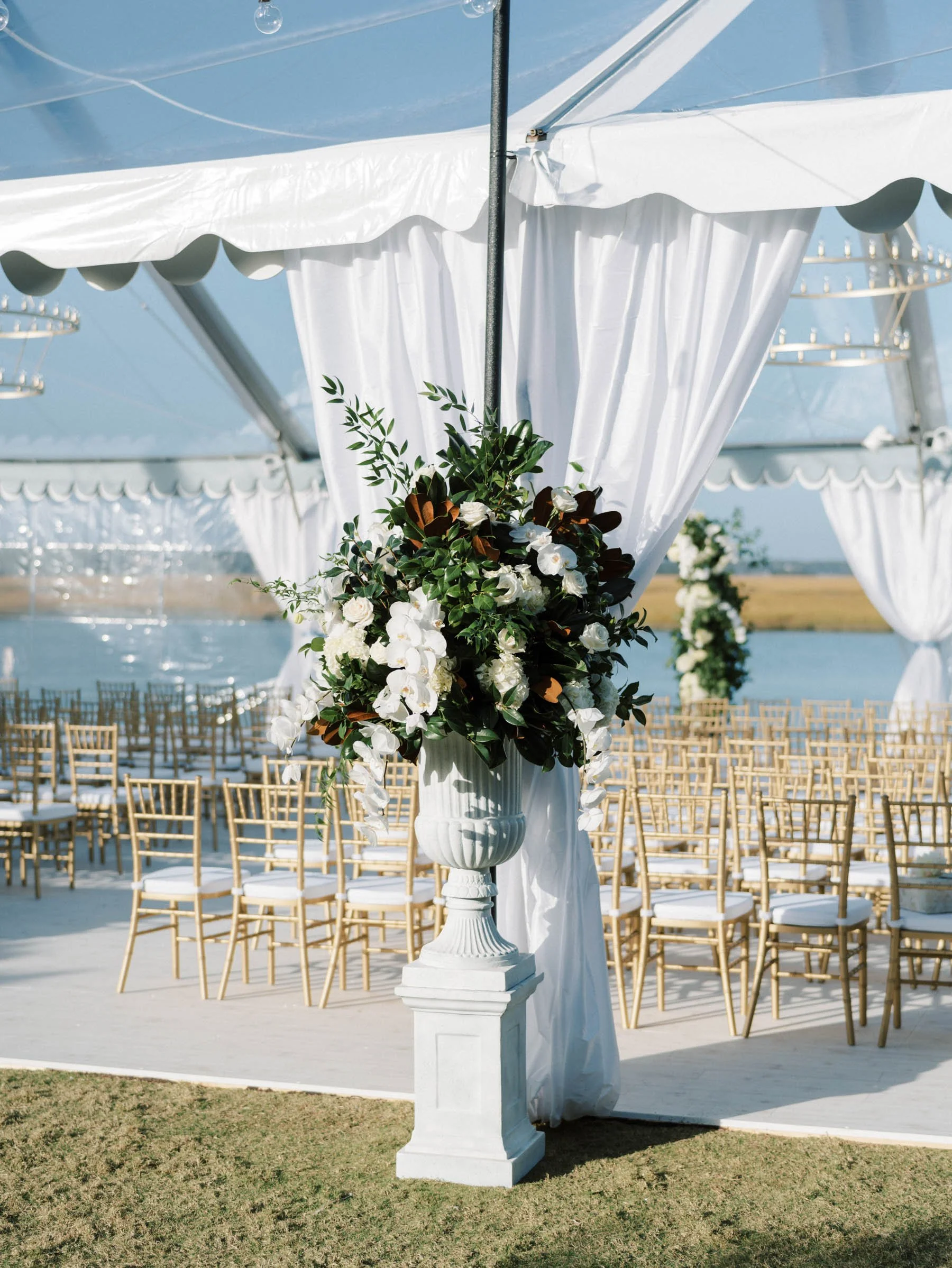 Outdoor wedding ceremony setup with white draped canopy, gold chairs, and floral arrangements on white pedestals near a body of water.