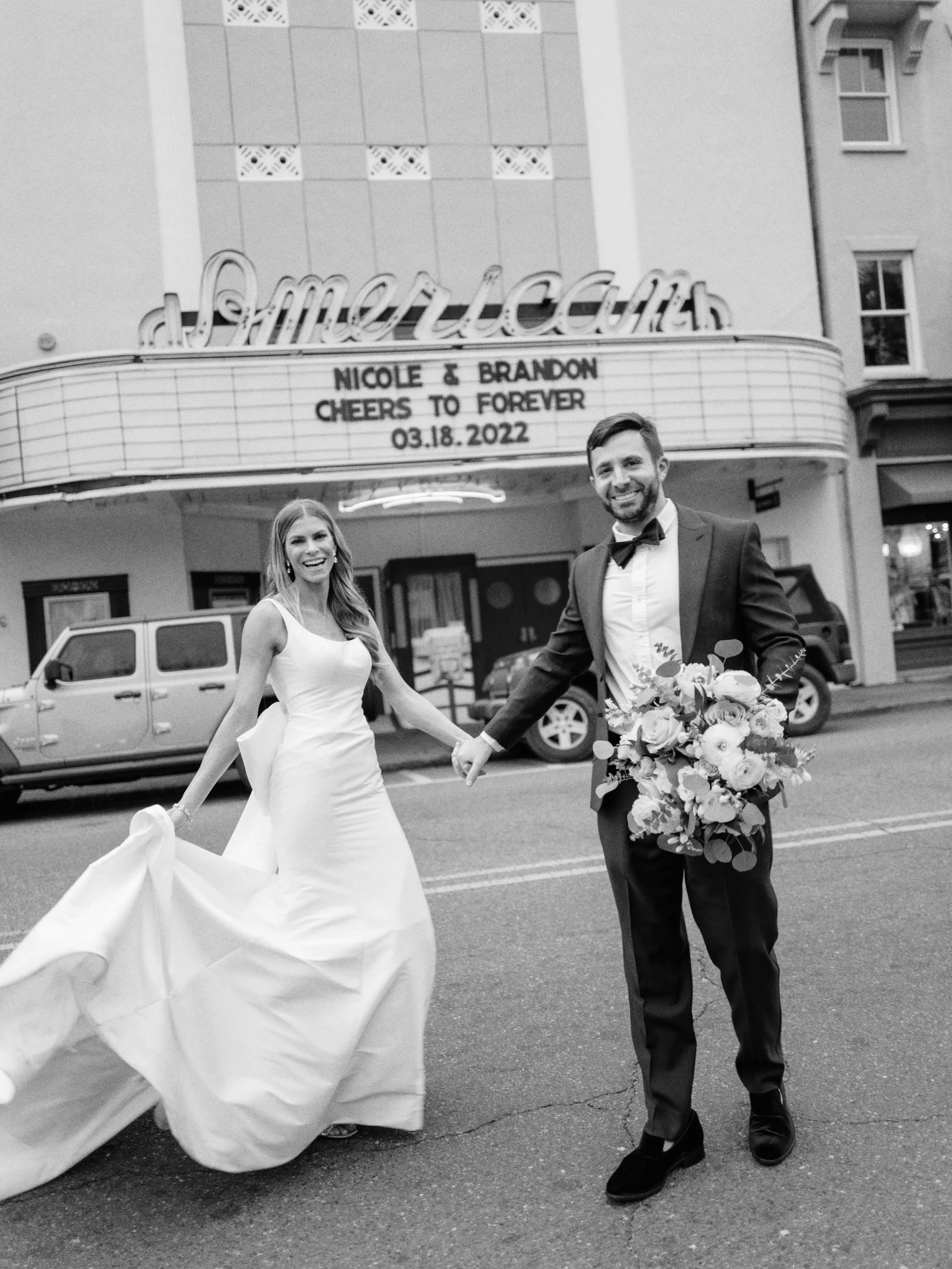 Black and white photo of a newlywed couple holding hands and smiling outside a theater marquee with the names Nicole and Brandon, date March 18, 2022, and a message 'Cheers to forever.' The bride is in a white wedding gown holding the train, and the groom in a tuxedo holding a bouquet of flowers.