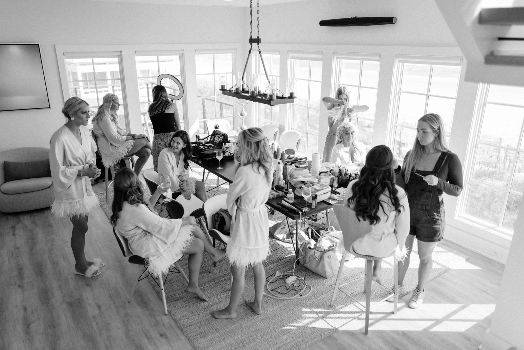 Black and white photo of a group of women in a bright modern room, some sitting and some standing, with makeup and hair styling tools on the table, preparing for an event.