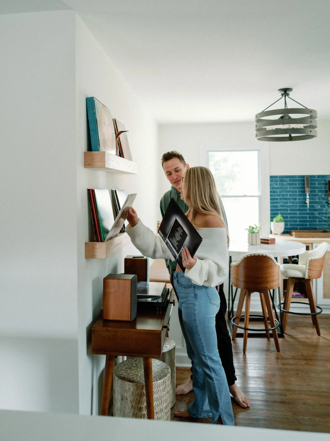 Two young adults, a man and a woman, are standing by a wall with shelves of books in a modern kitchen, looking at a tablet and a framed picture.