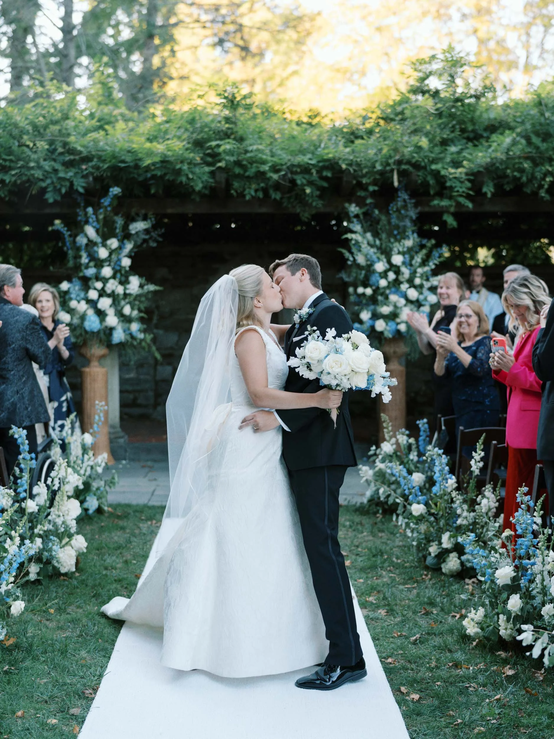 A bride and groom kiss during their outdoor wedding ceremony. The bride wears a white wedding gown with a veil, and the groom is in a black tuxedo. They are surrounded by friends and family, with floral arrangements and greenery in the background.