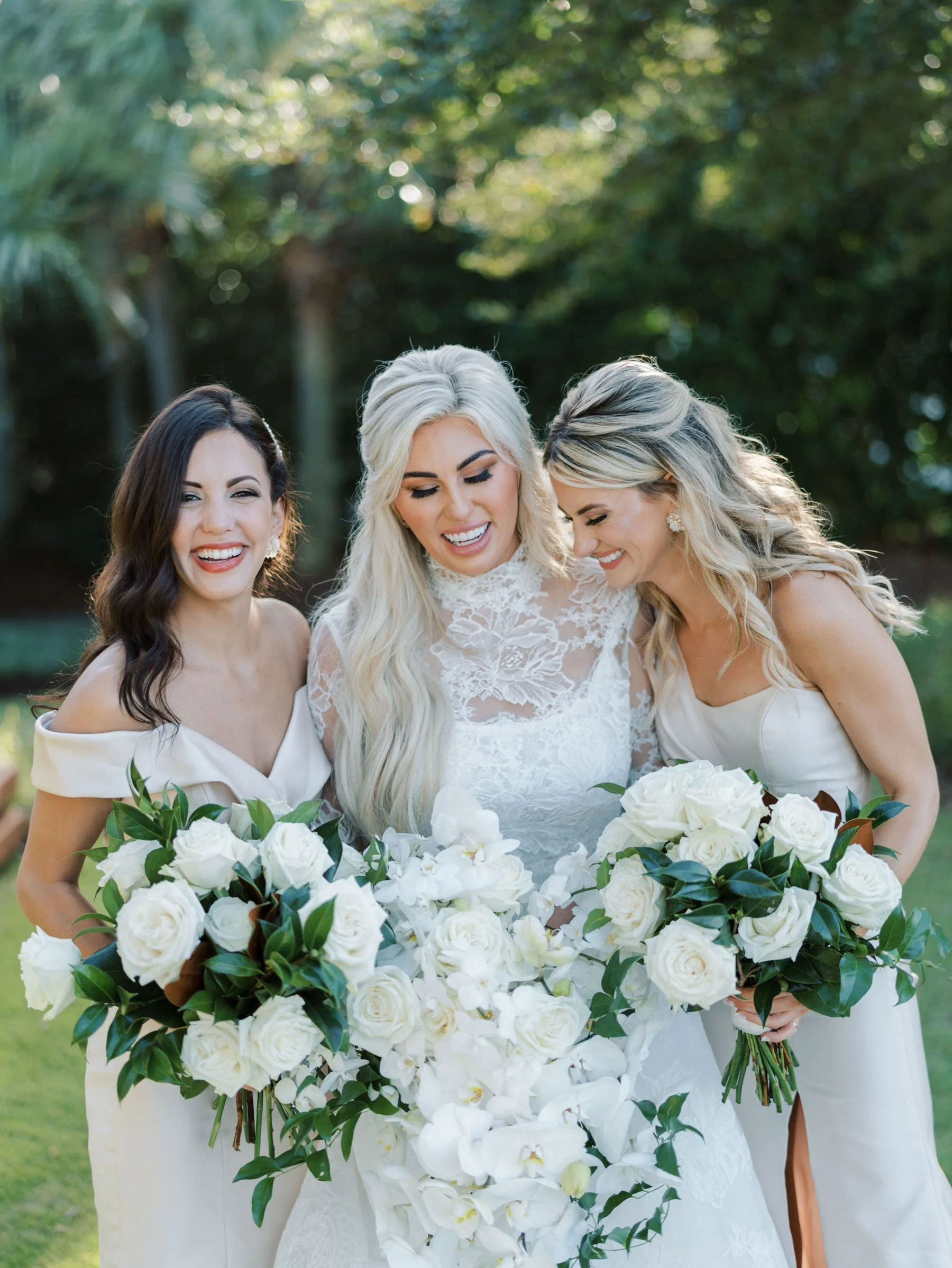 Three women in wedding dresses holding large bouquets of white roses and orchids, smiling and enjoying the moment outdoors with trees in the background.