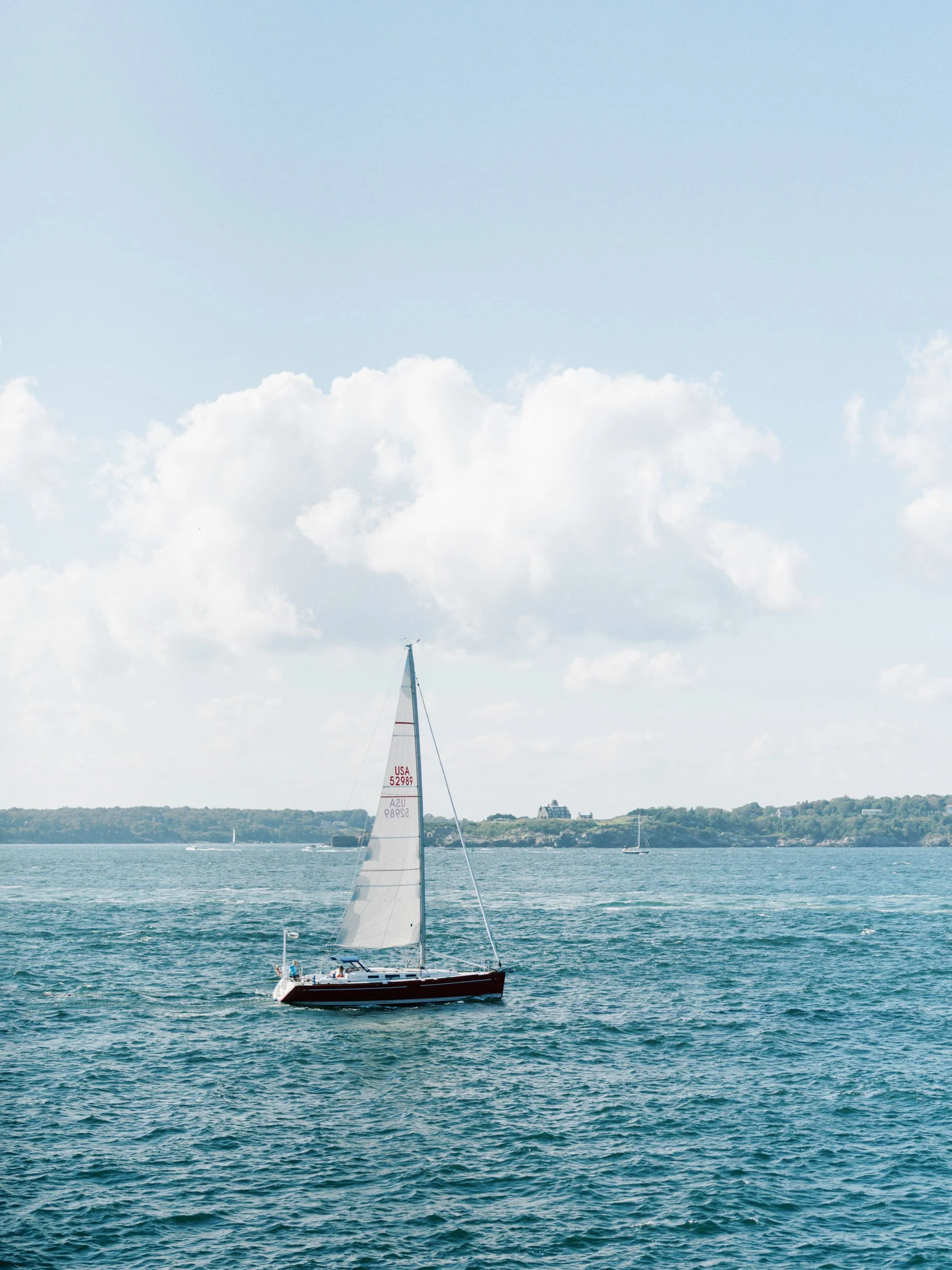 A sailboat with white sails on a large body of water, with land and houses in the distance under a partly cloudy sky.