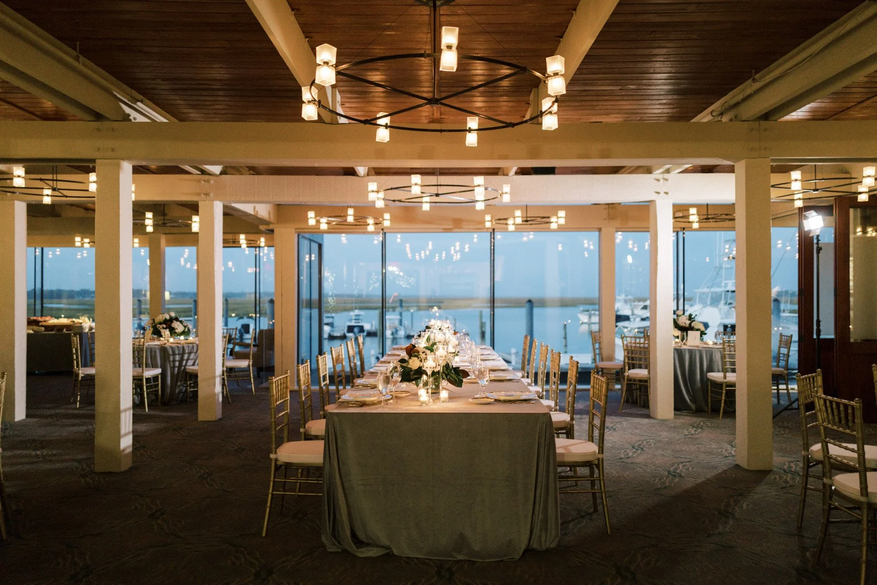Elegant dining setup with a long table decorated with flowers, candles, and tableware, overlooking a marina through large glass windows, illuminated by hanging ceiling lights.