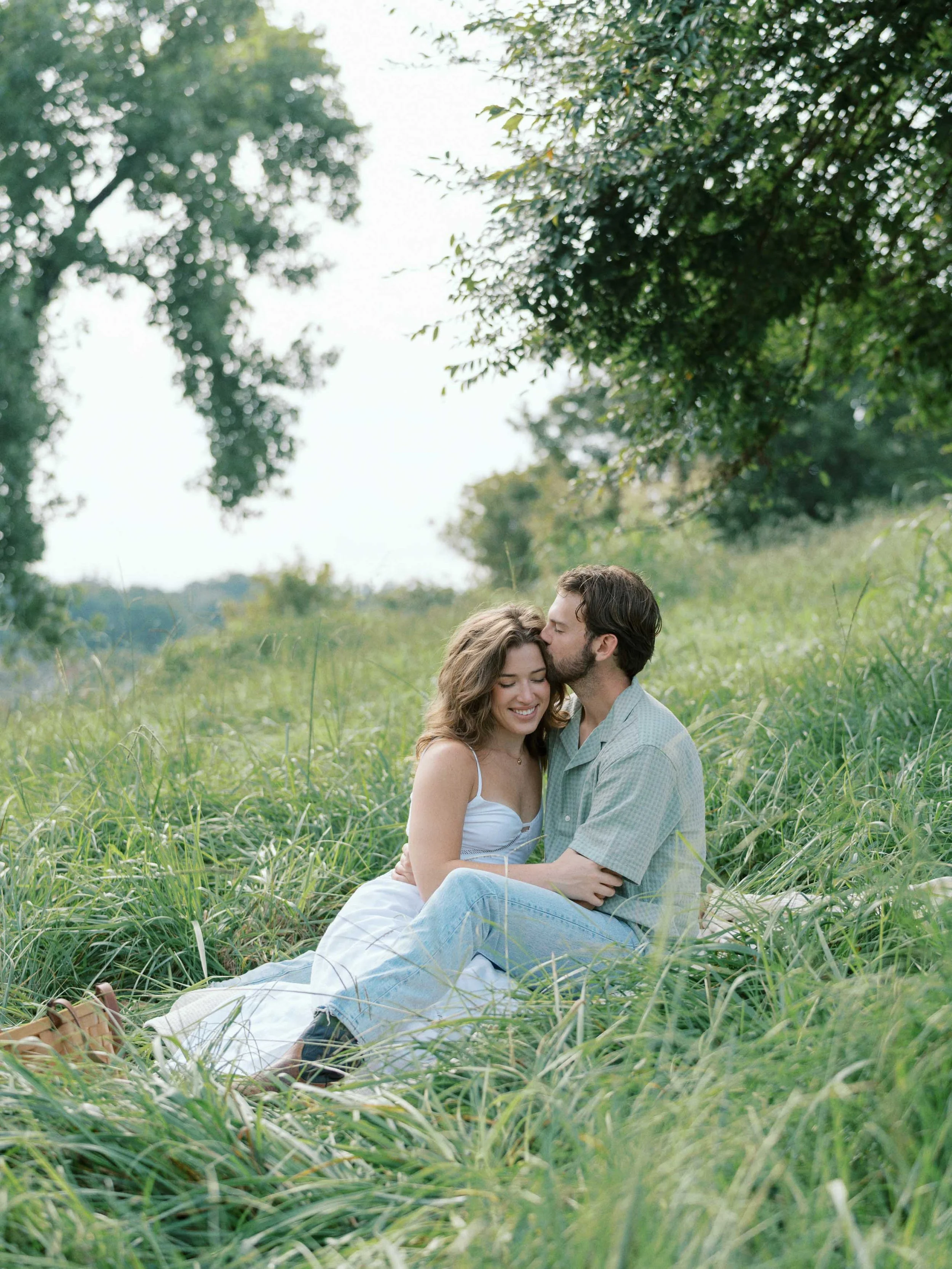 A couple sitting together in a grassy field, with the man kissing the woman's forehead and both smiling, under the shade of a large tree with a scenic background.