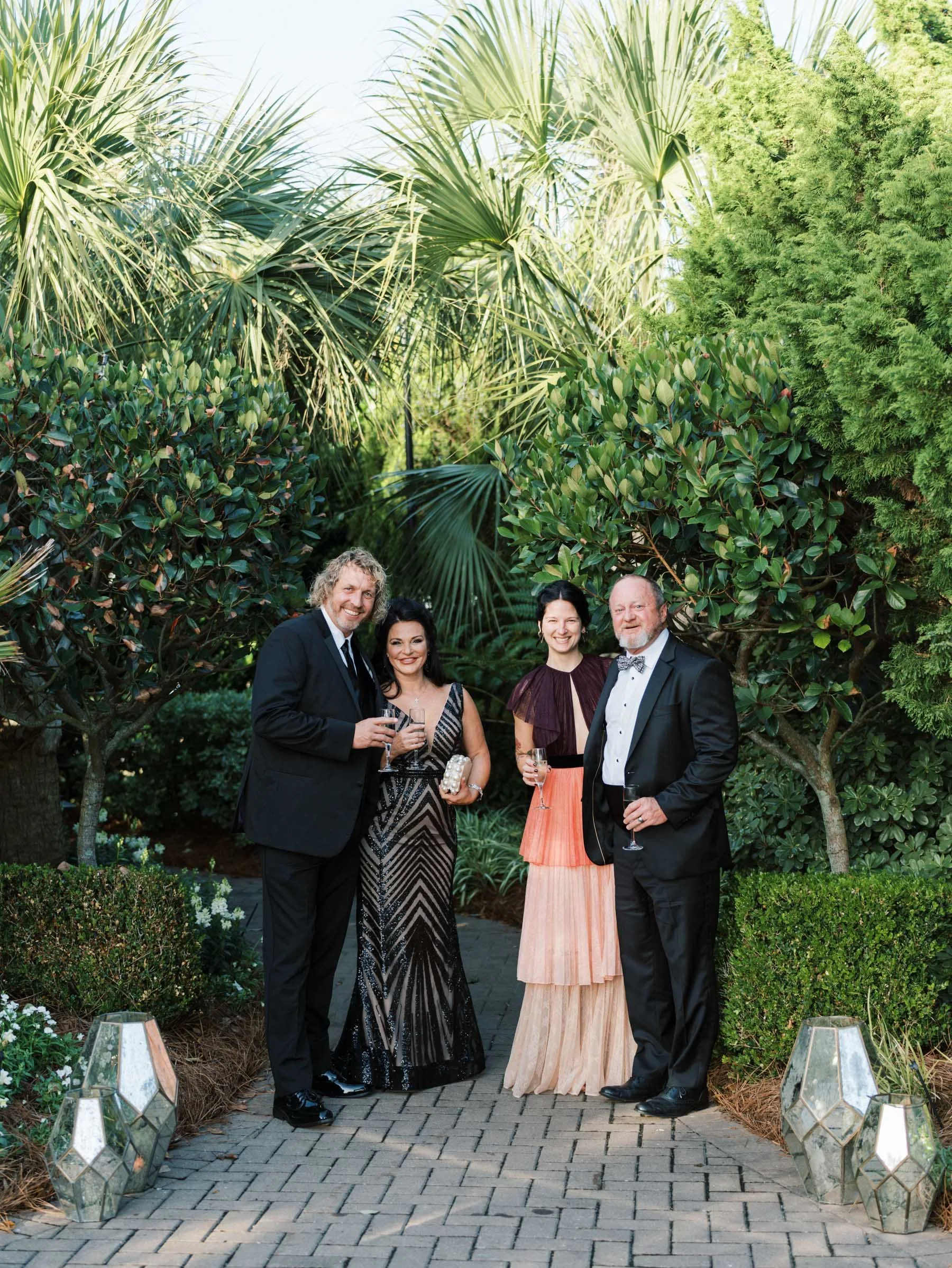 Group of four people in formal attire standing on a brick pathway surrounded by lush greenery, holding glasses of champagne, at an outdoor event.