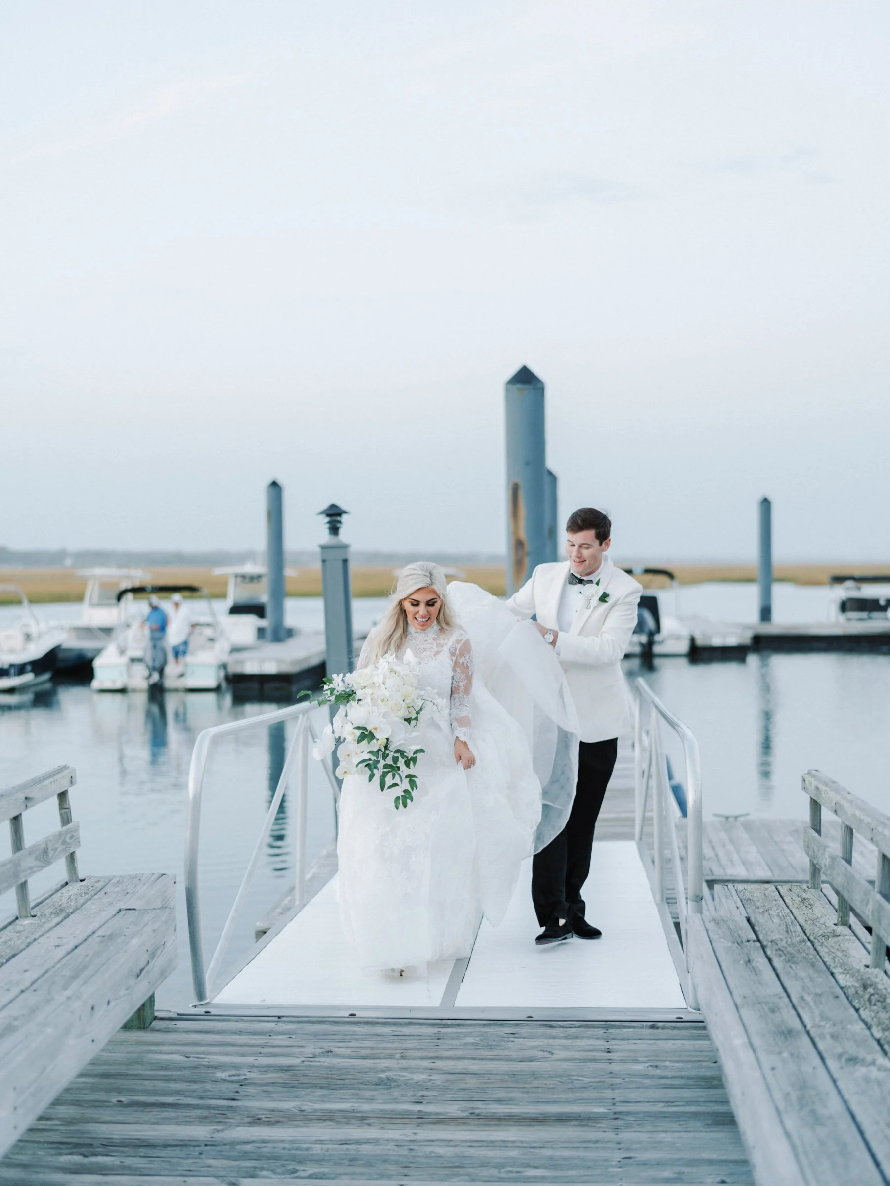 Bride and groom walking on a dock by the water, with boats docked in the background, during their wedding celebration.