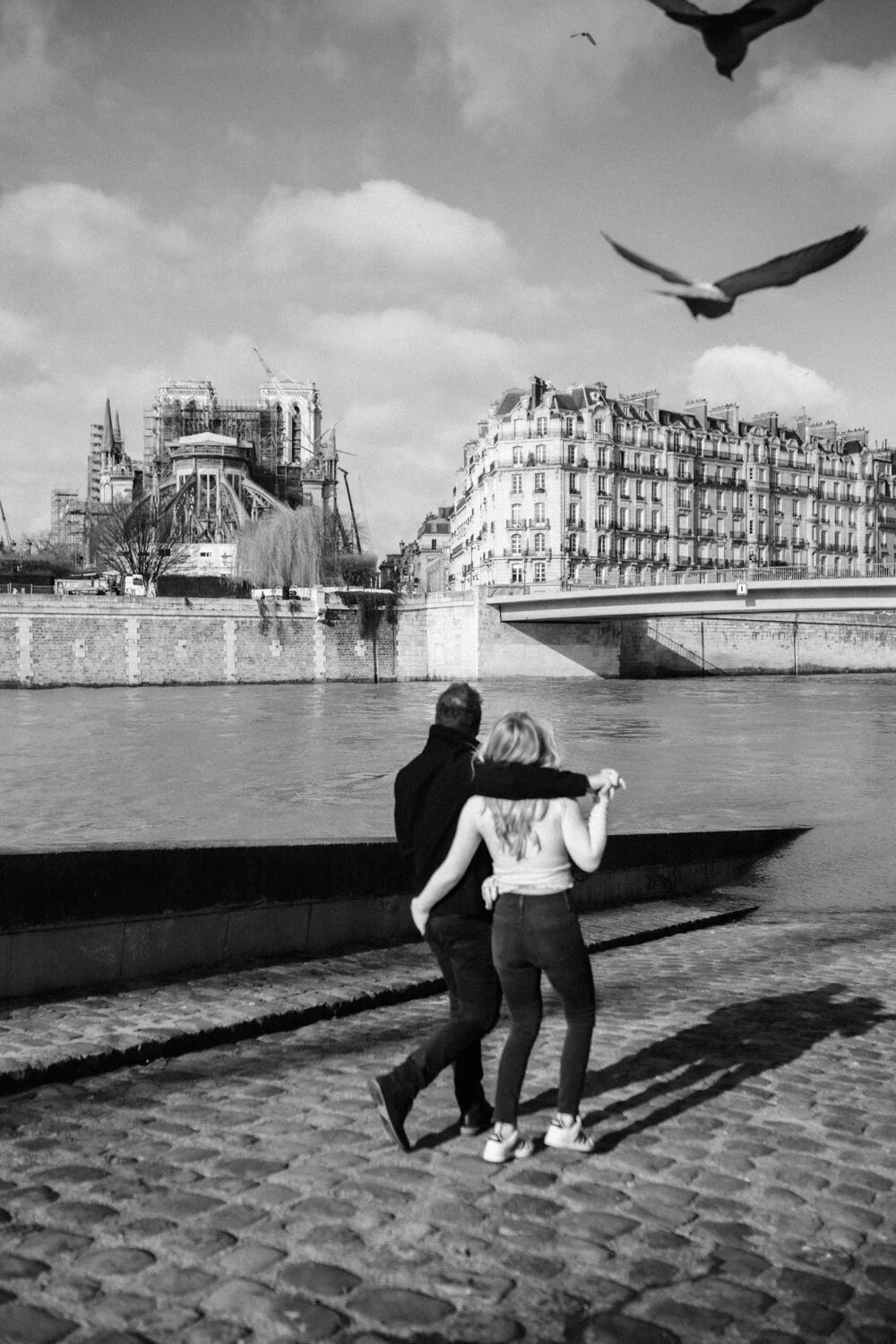 A couple walking along a riverside in Paris, with the Eiffel Tower under construction and several birds flying overhead.