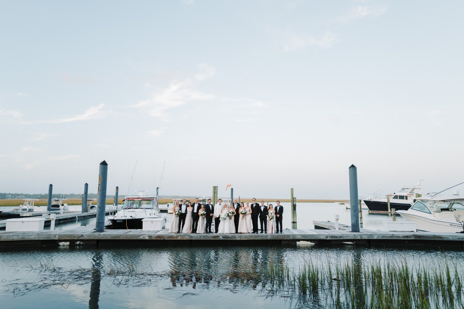 A wedding party standing on a dock by the water with boats in the background, under a partly cloudy sky.