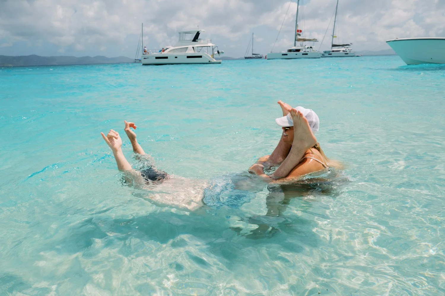 Two women in a clear, turquoise ocean embracing and hugging each other, with sailboats and yachts anchored in the background under a partly cloudy sky.
